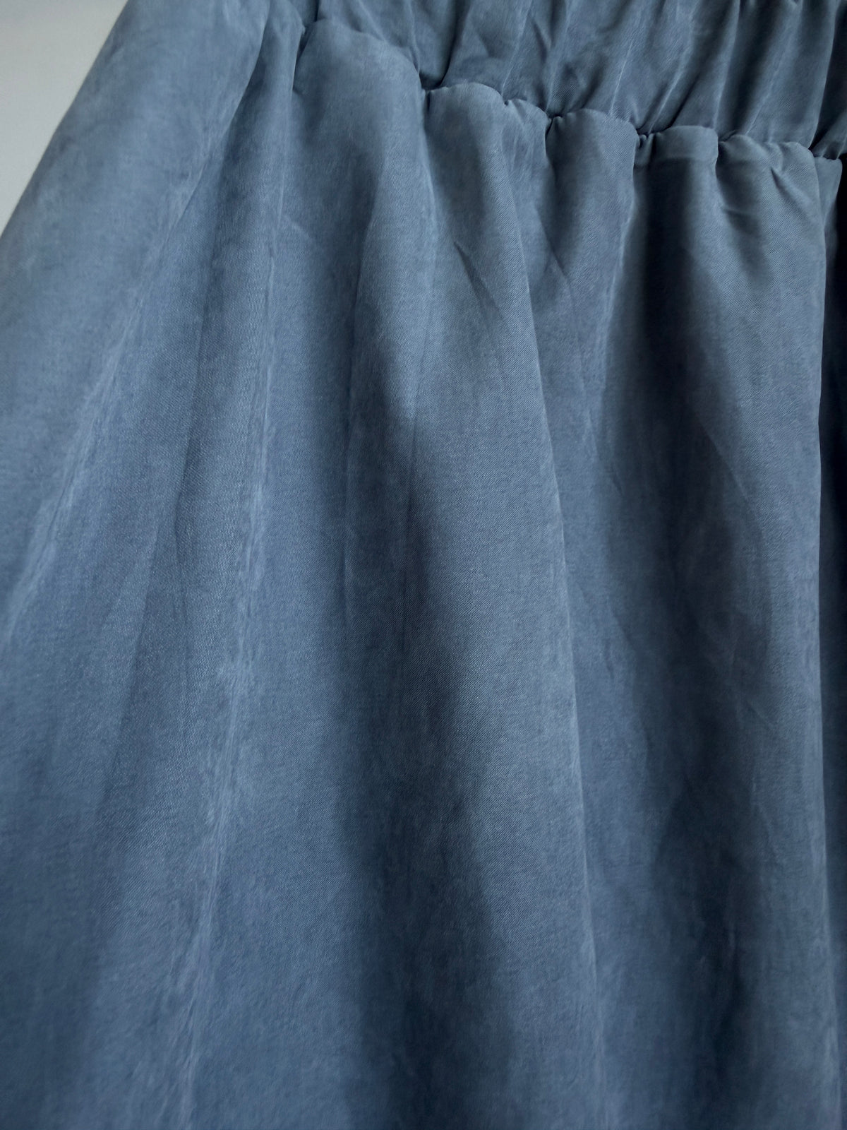 A close-up of a dusty slate blue fabric, showing the texture and gathered waist seam of a garment.