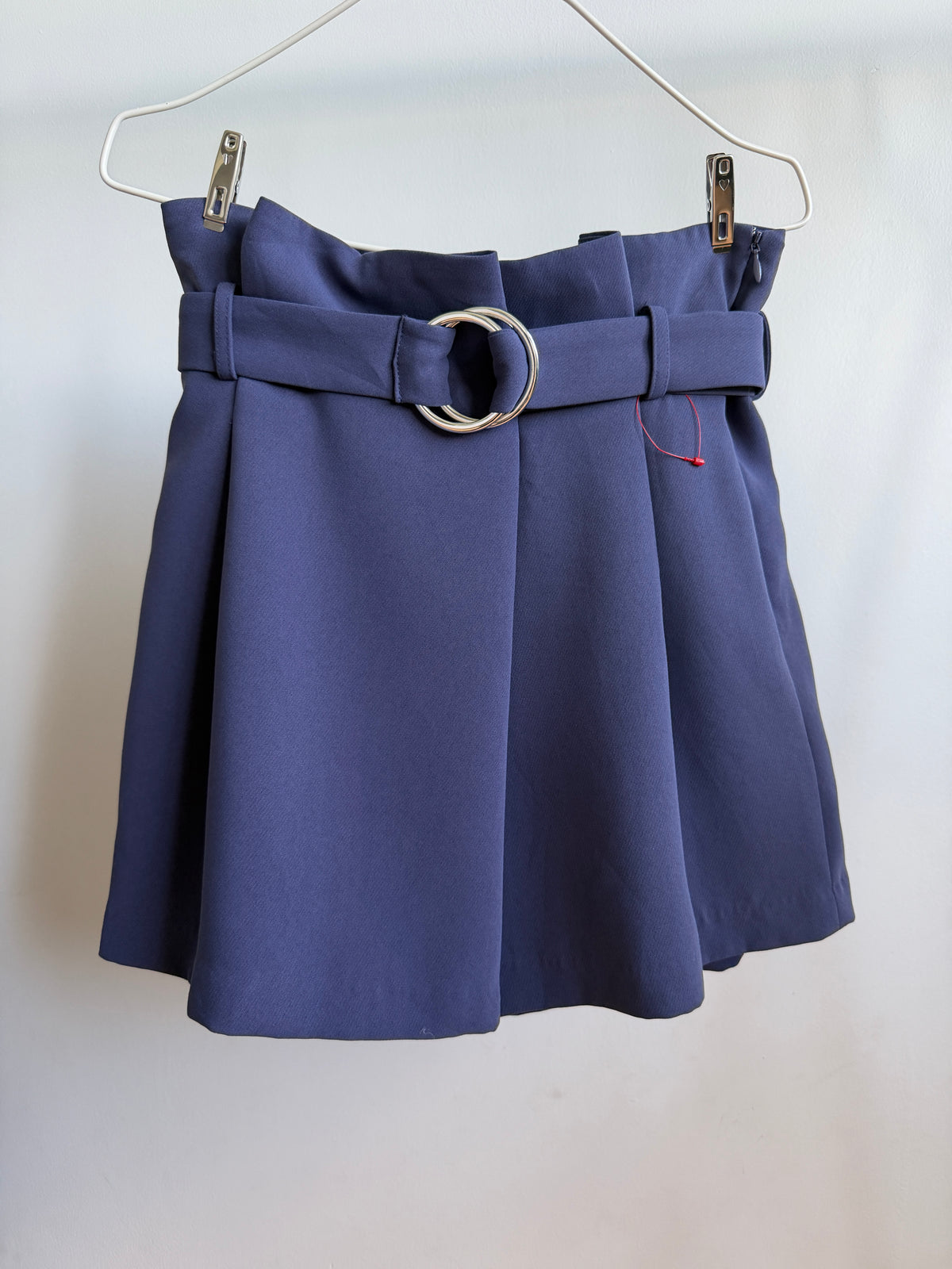A navy blue pleated mini skirt with a matching waist belt and a large silver-tone double-ring buckle, hanging from a metal wire hanger against a plain off-white background.