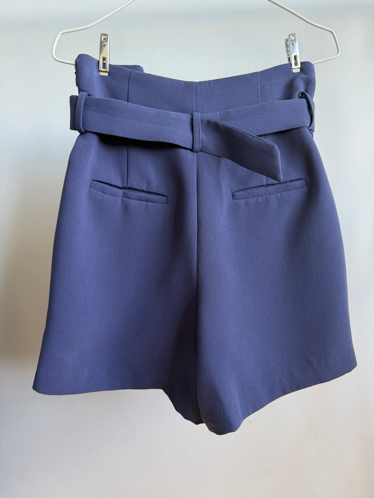 A pair of high-waisted, wide-leg shorts in a dusty periwinkle blue, hanging from a white wire hanger with silver clips against a plain off-white wall. The shorts feature a matching fabric tie belt at the waist, two front welt pockets, and a structured silhouette.