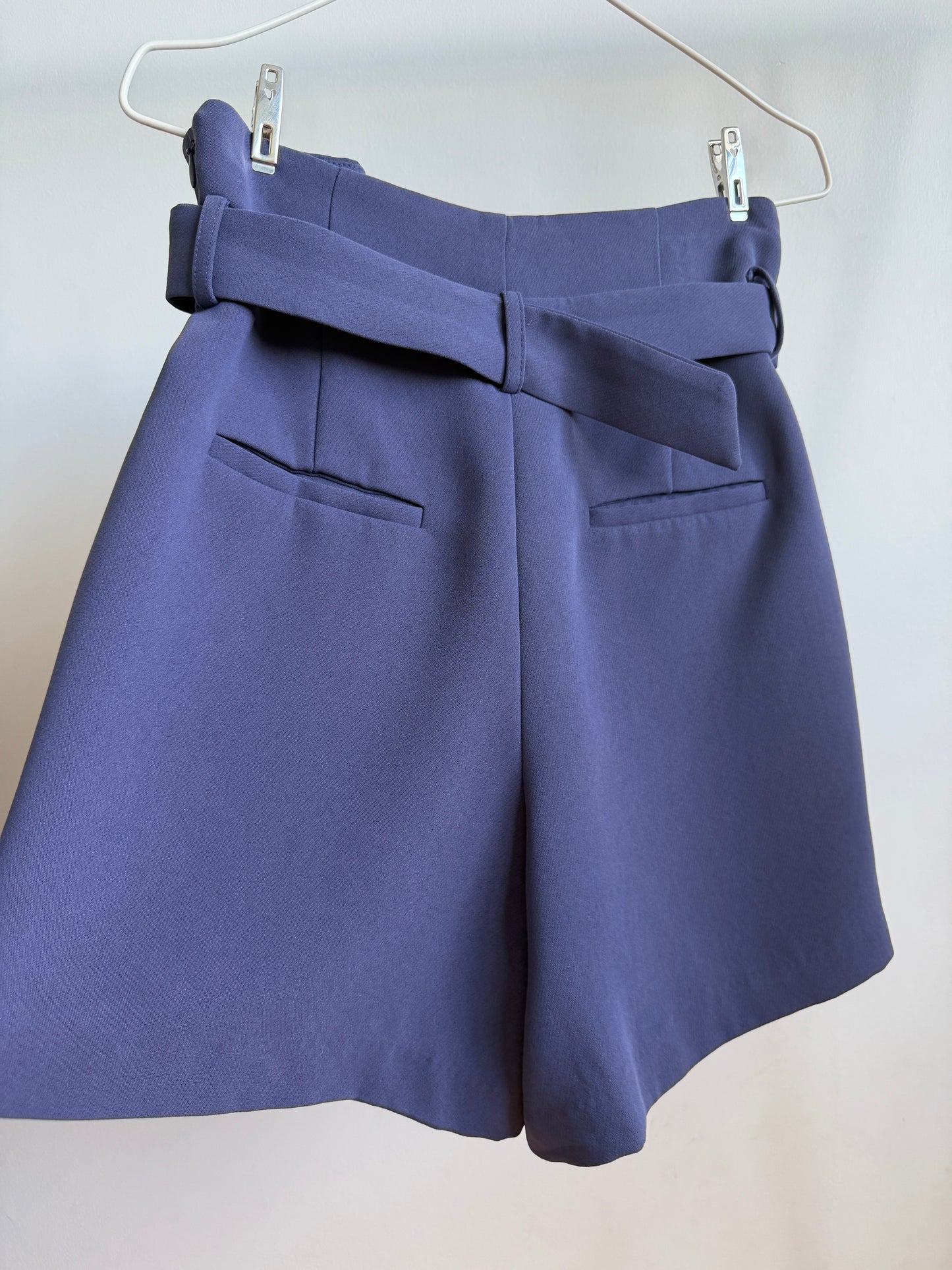 A pair of high-waisted, wide-leg shorts in a dusty periwinkle blue-purple color, hanging from a white wire hanger with metal clips against a plain off-white wall.
