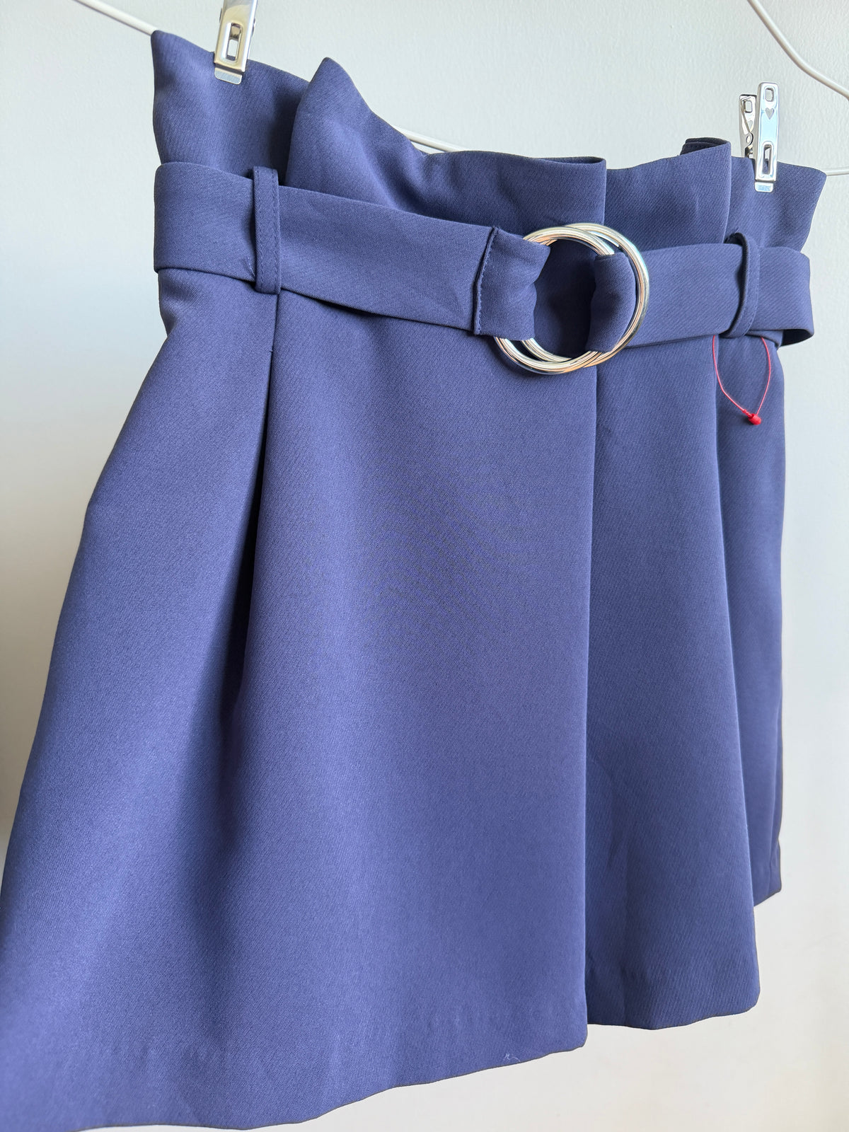 A high-waisted blue mini skirt with a matching waist belt and a large silver-tone double-ring buckle, hanging on a white hanger against a plain light gray wall.
