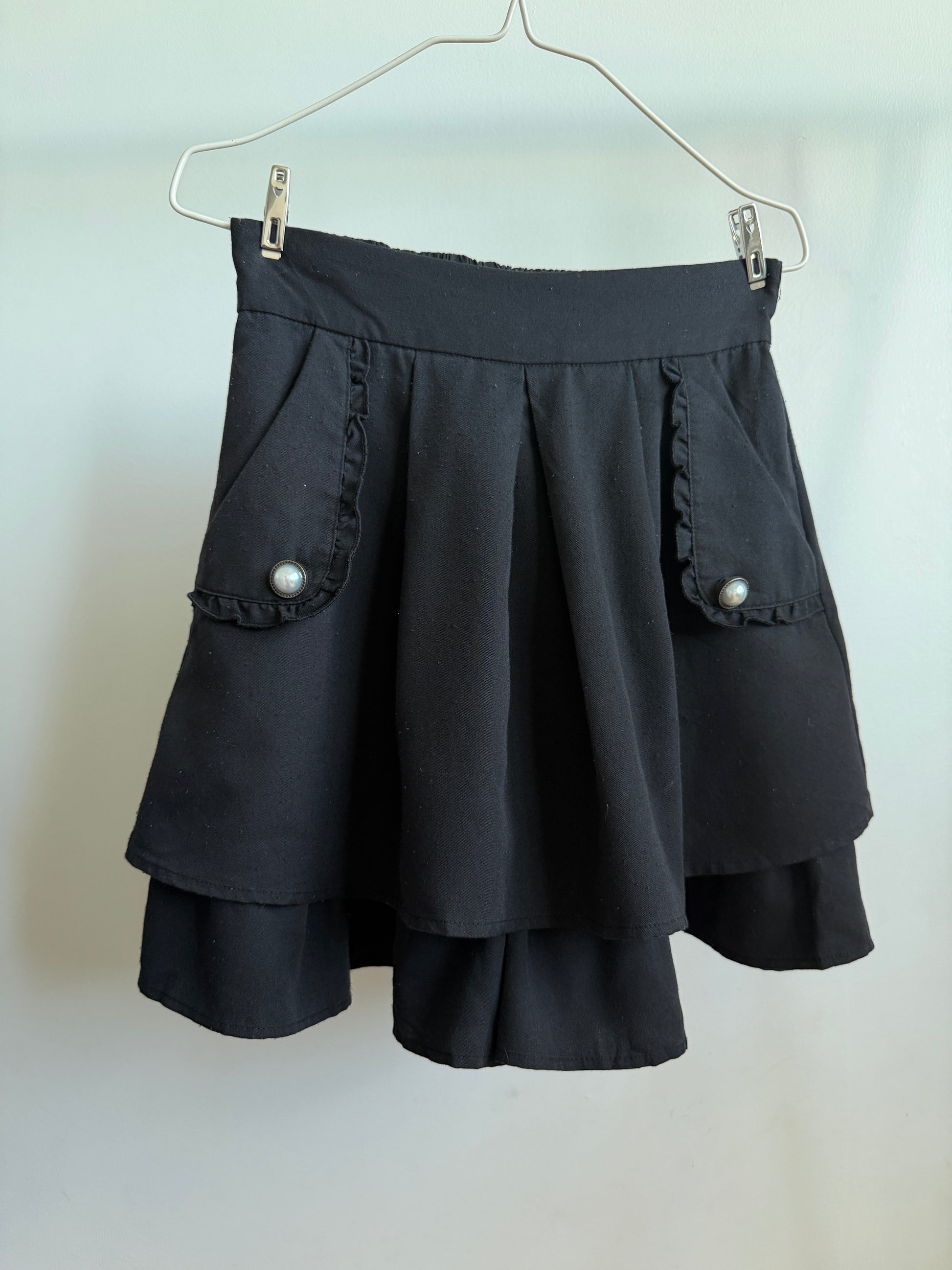 A black pleated mini skirt with ruffled pocket trim and pearl-like button details, hanging from a metal clothes hanger against a plain light background.