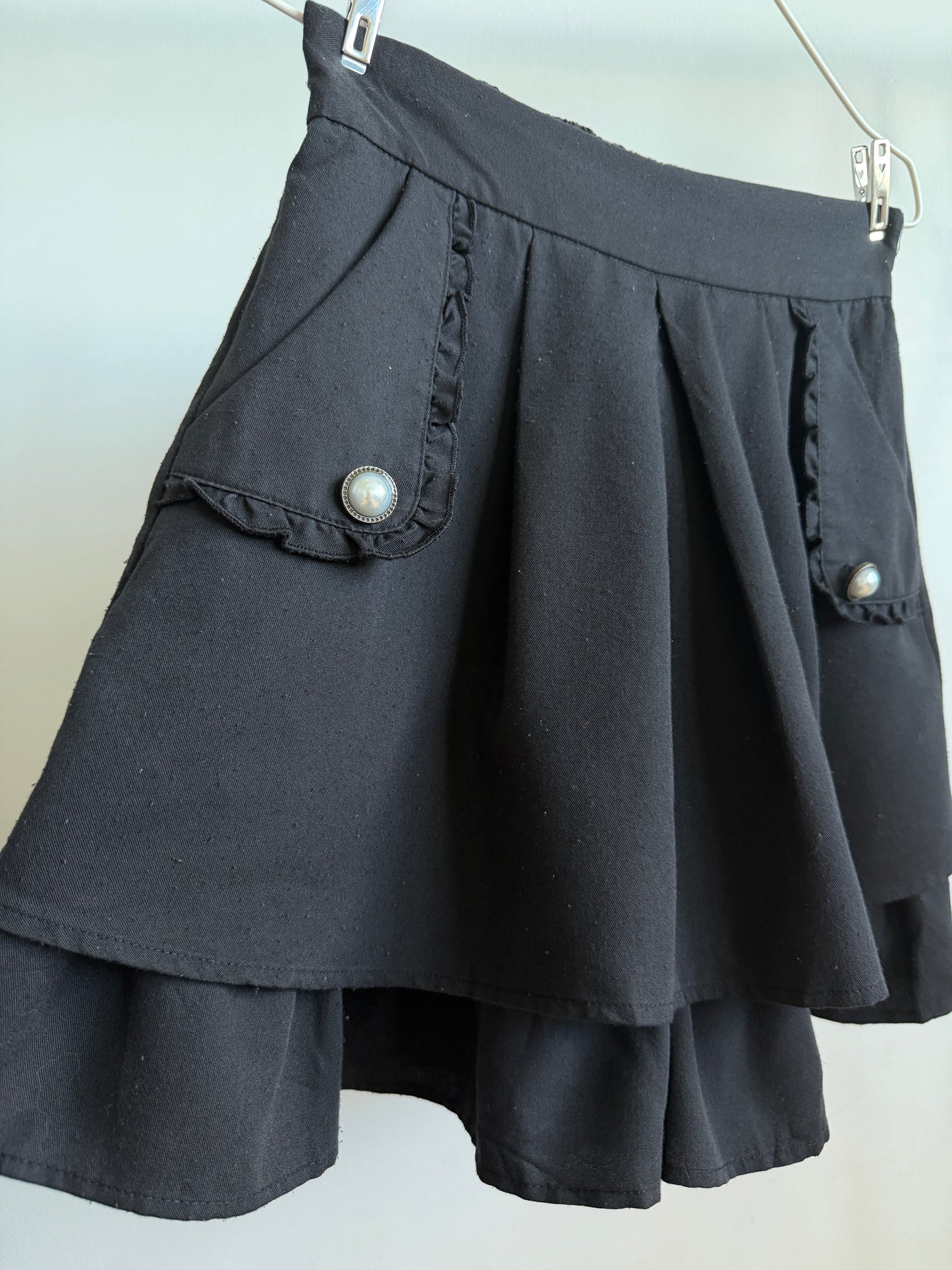 A black pleated mini skirt with ruffled pocket flaps and decorative pearl-like buttons, hanging against a light background.