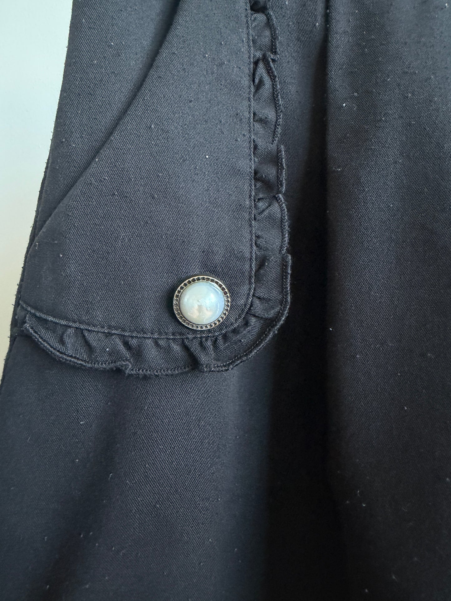 Close-up of a black fabric garment featuring a ruffled pocket flap with a decorative round button.