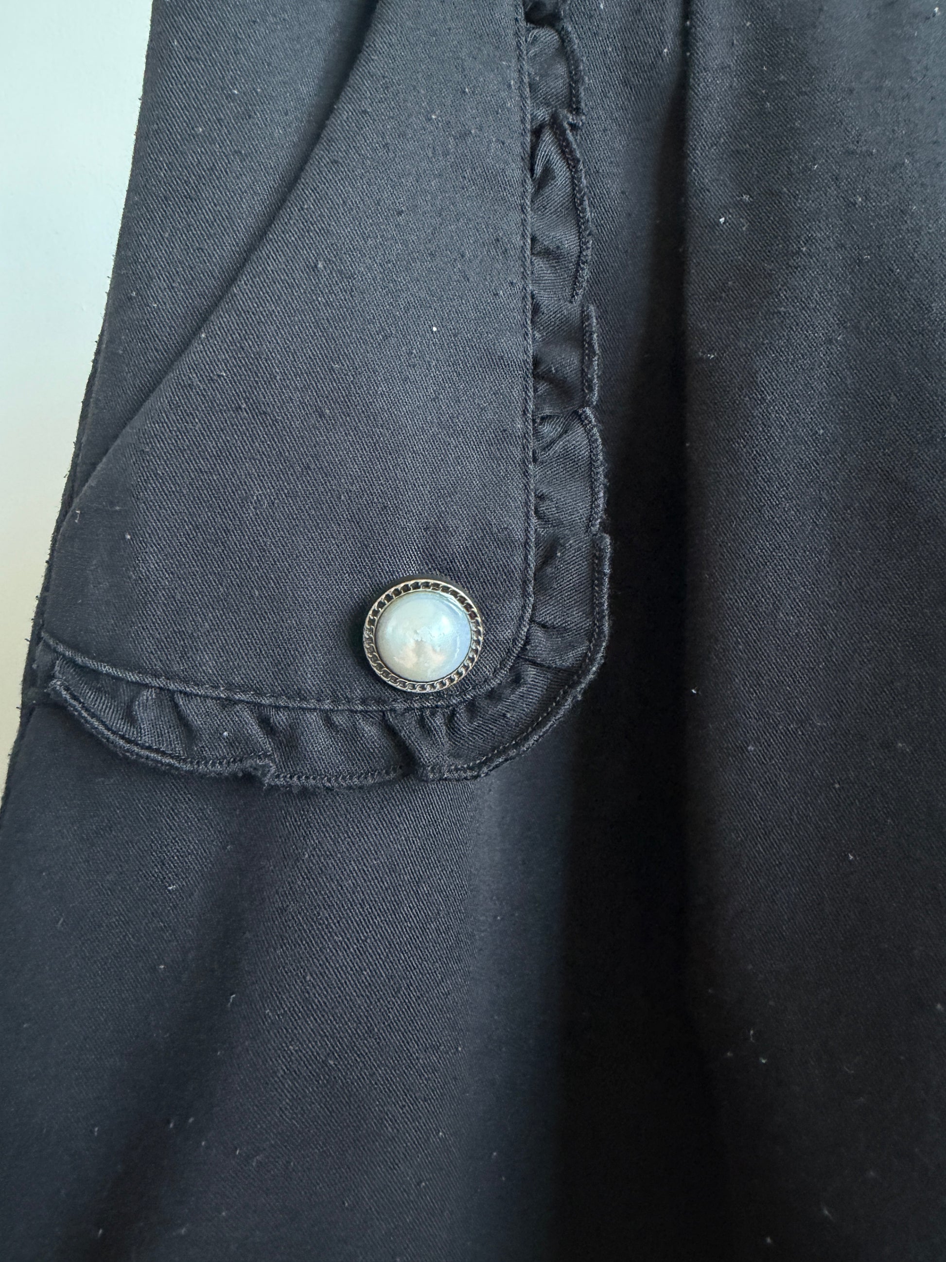 Close-up of a black fabric garment featuring a ruffled pocket flap with a decorative round button.