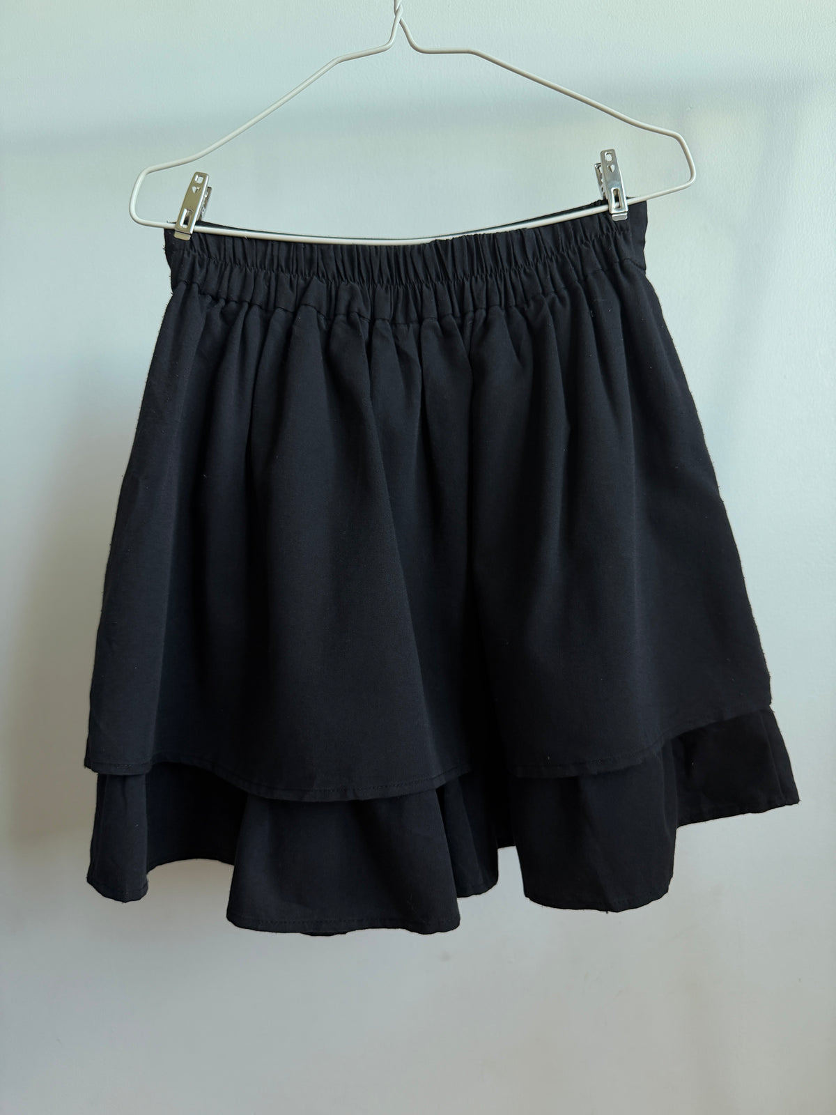 A black tiered mini skirt with an elastic waistband, hanging from a metal wire hanger against a plain light gray wall.