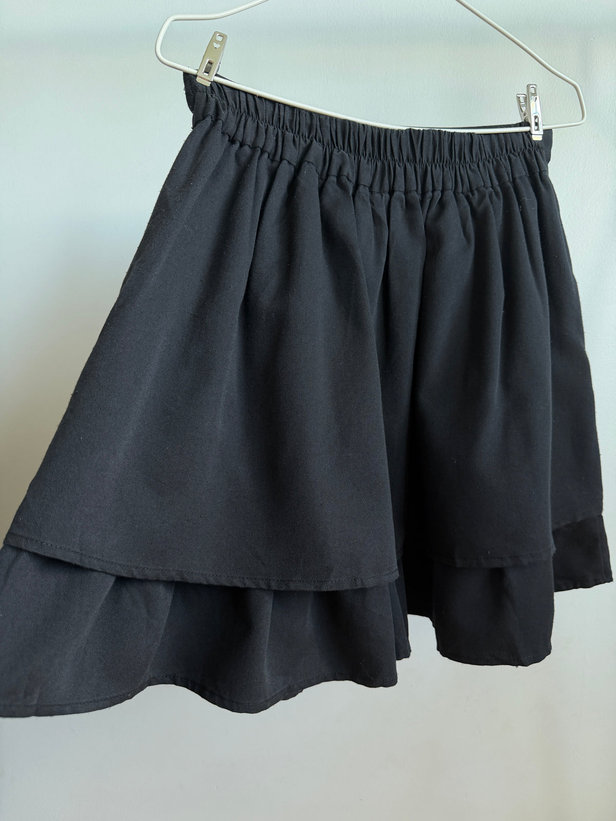 A black tiered skirt with an elastic waistband, hanging from a white hanger against a plain light gray wall.
