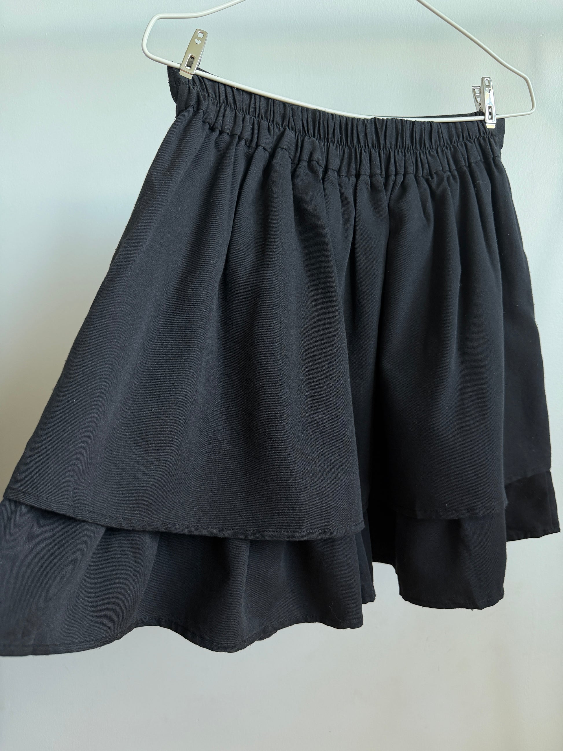 A black tiered skirt with an elastic waistband, hanging from a white hanger against a plain light gray wall.