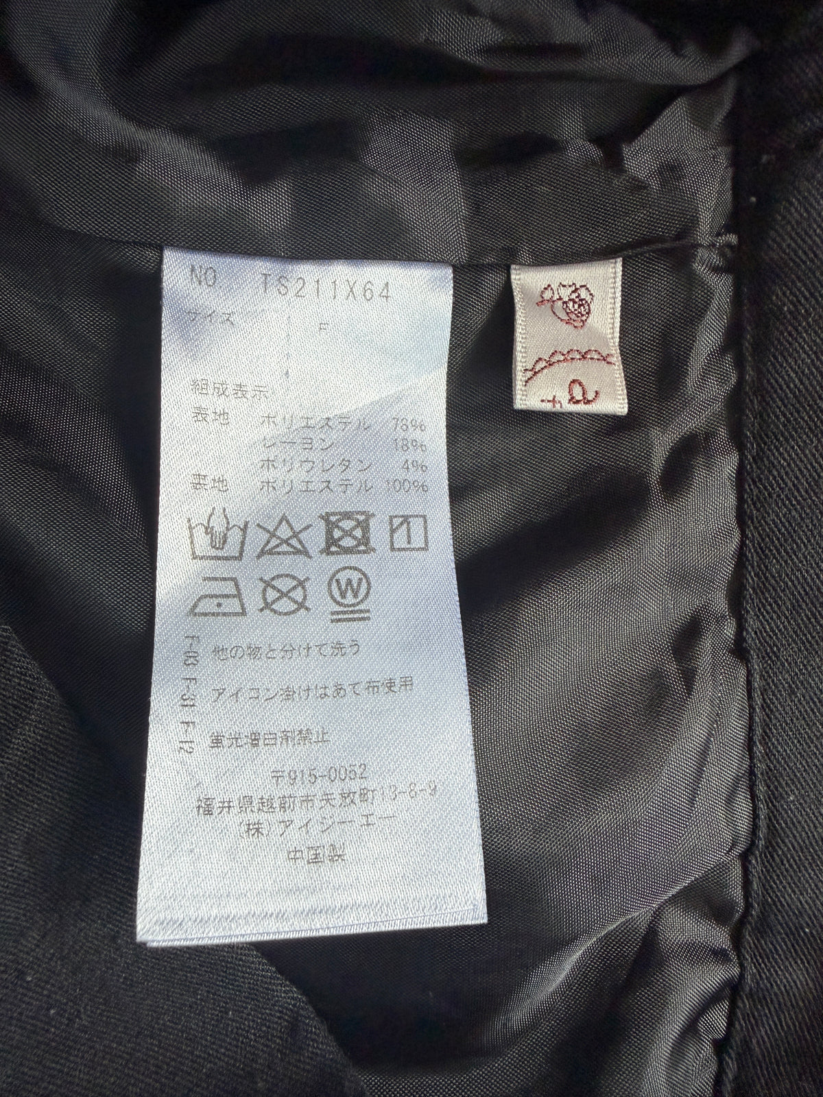 Close-up of the interior collar of a black garment showing a white size and care label with Japanese text, plus a small sewn brand tag with a red embroidered symbol.