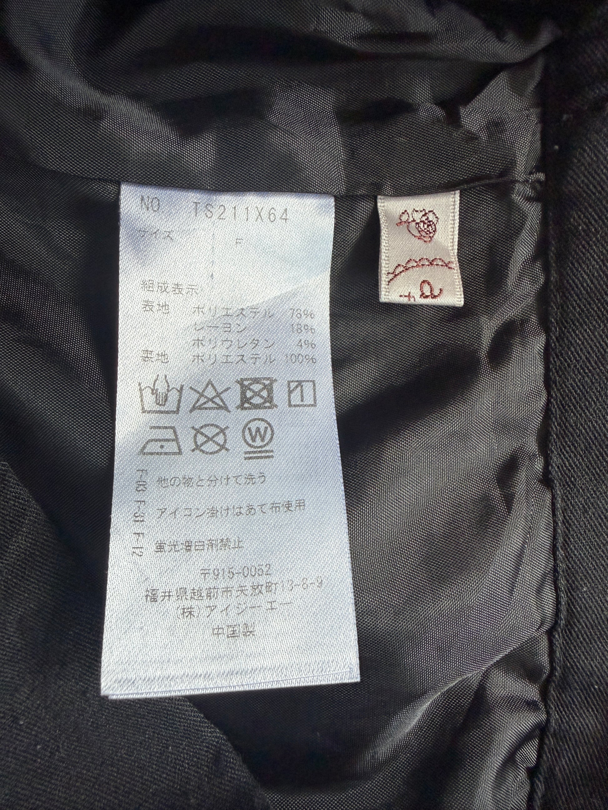 Close-up of the interior collar of a black garment showing a white size and care label with Japanese text, plus a small sewn brand tag with a red embroidered symbol.