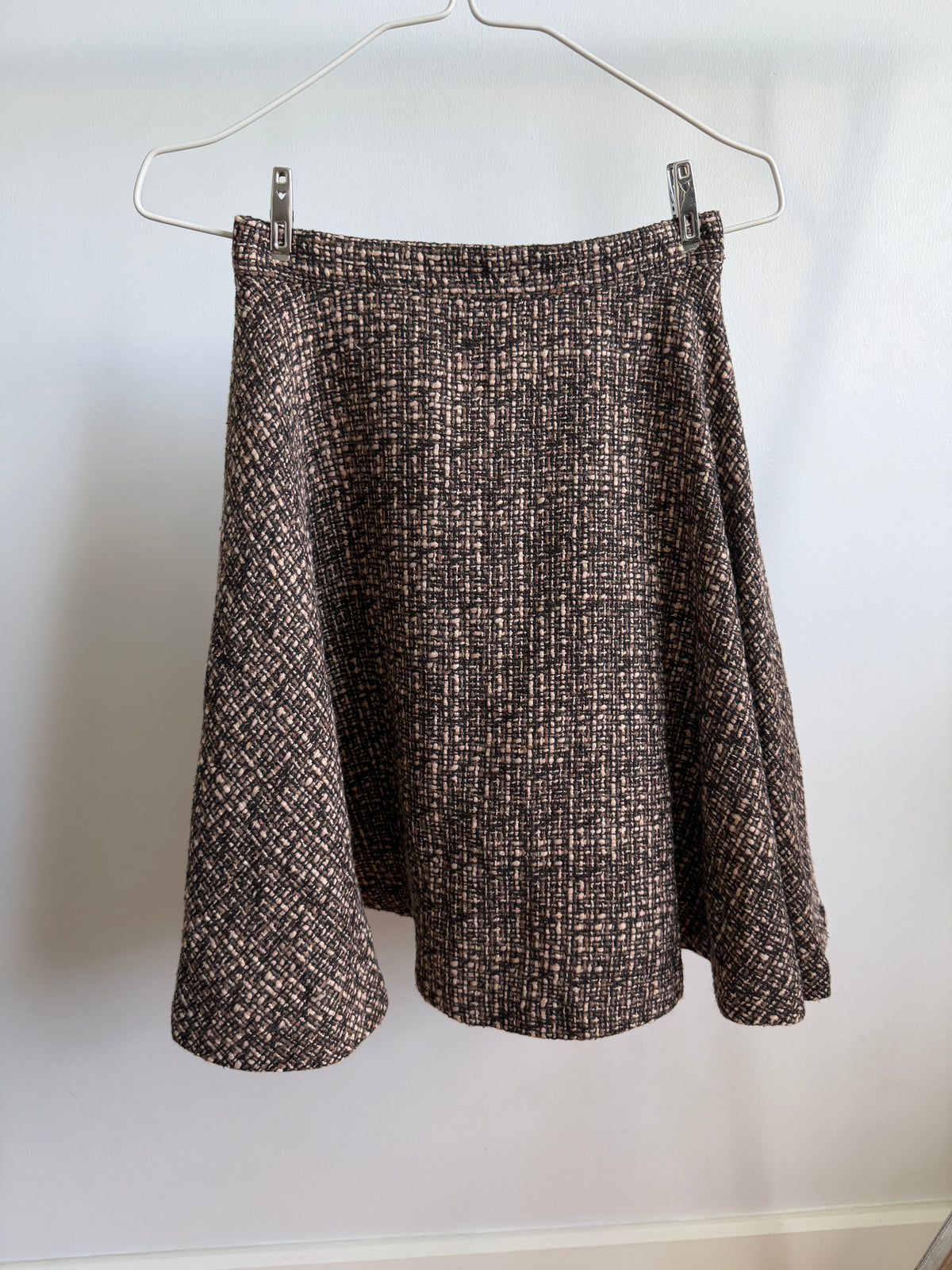 A brown and beige textured tweed skirt hanging from a metal hanger against a plain white wall.