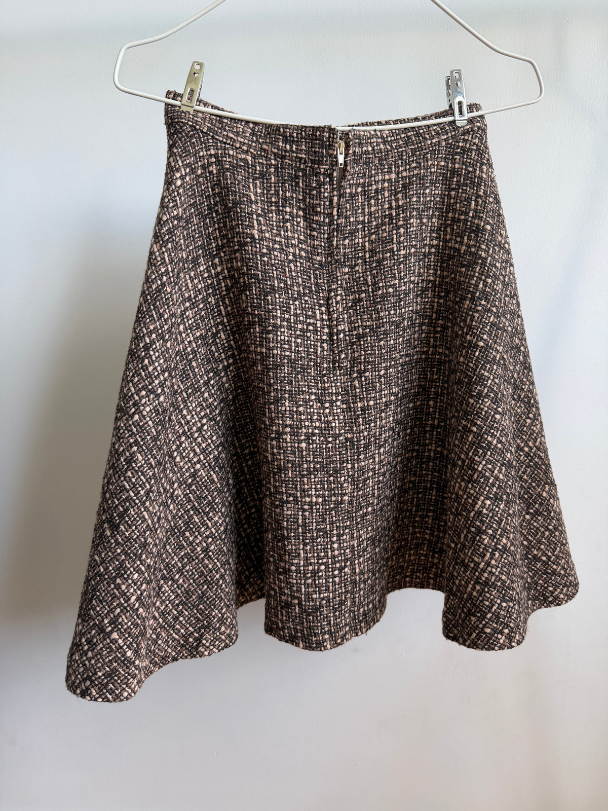 A brown and beige tweed A-line skirt hanging from a white wire hanger against a plain light gray wall.