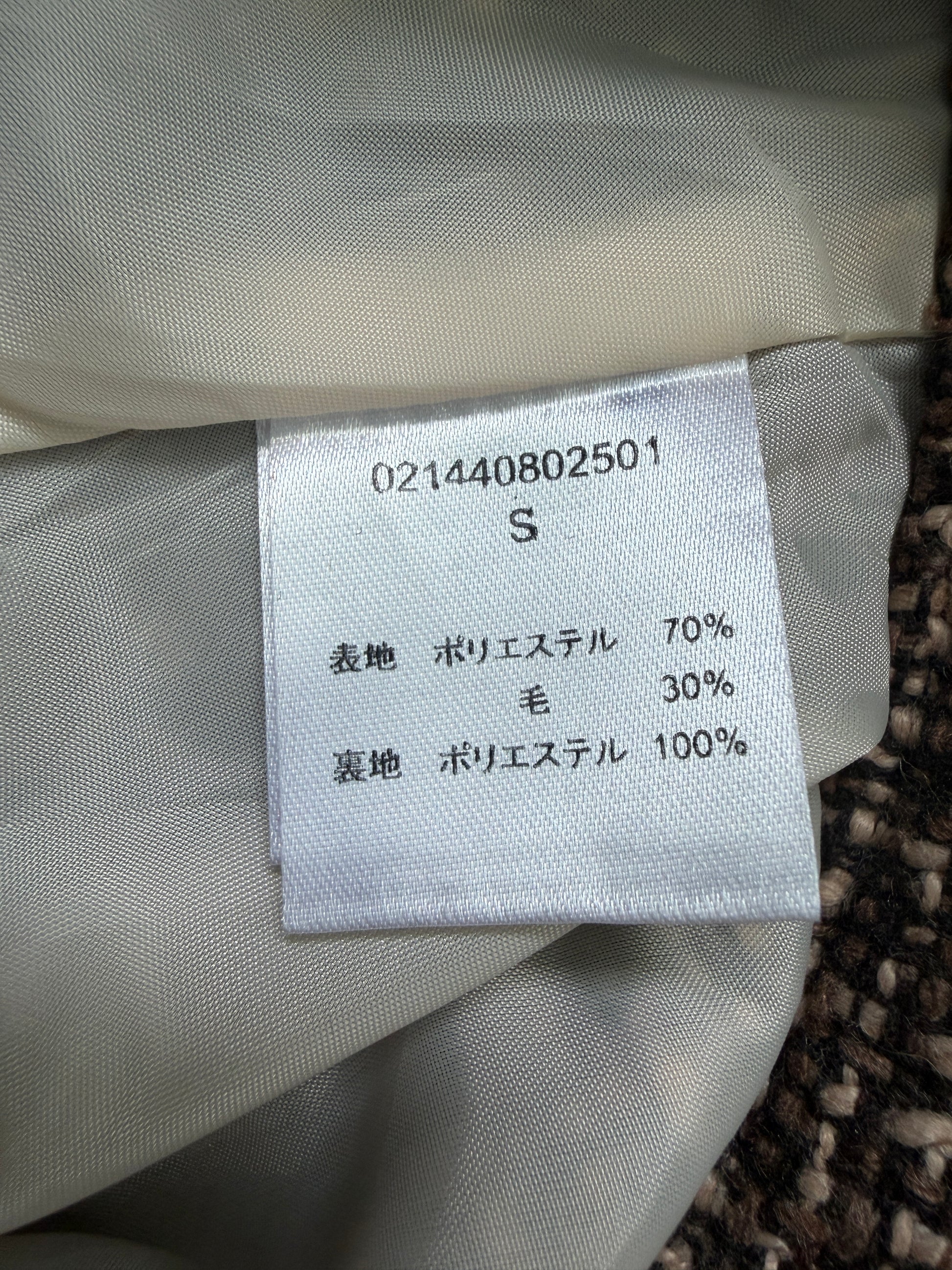 Close-up of a garment's interior size label reading 'S' with material composition details in Japanese, attached to light beige fabric.