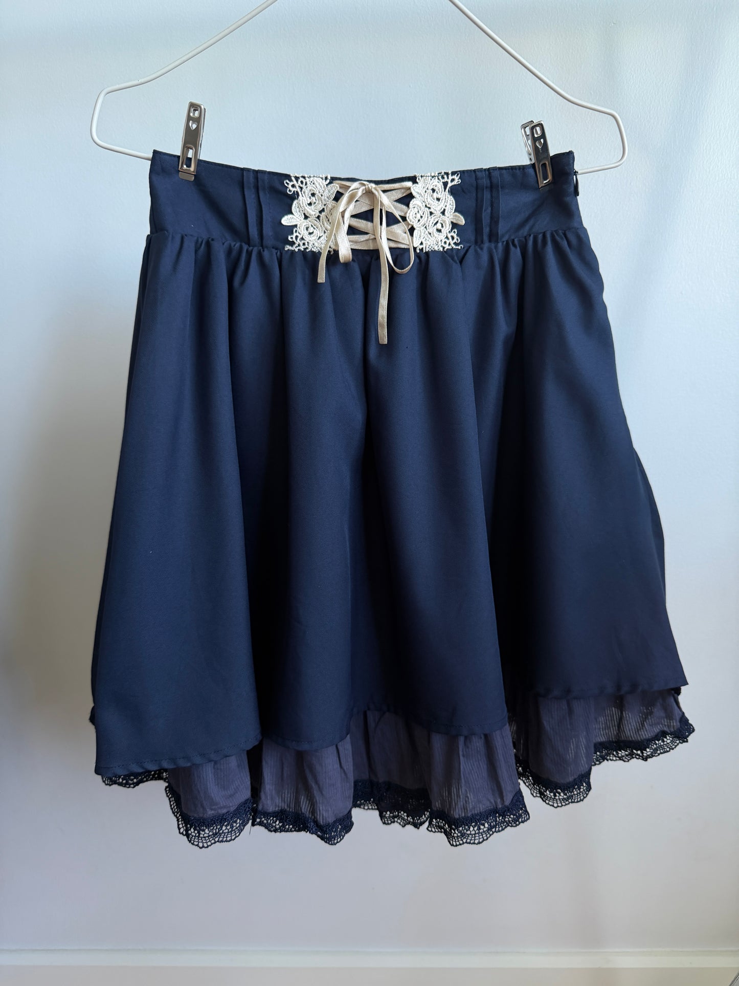 A navy blue skirt with a lace-up waistband and white floral lace detail, hanging from a metal hanger against a plain light background.