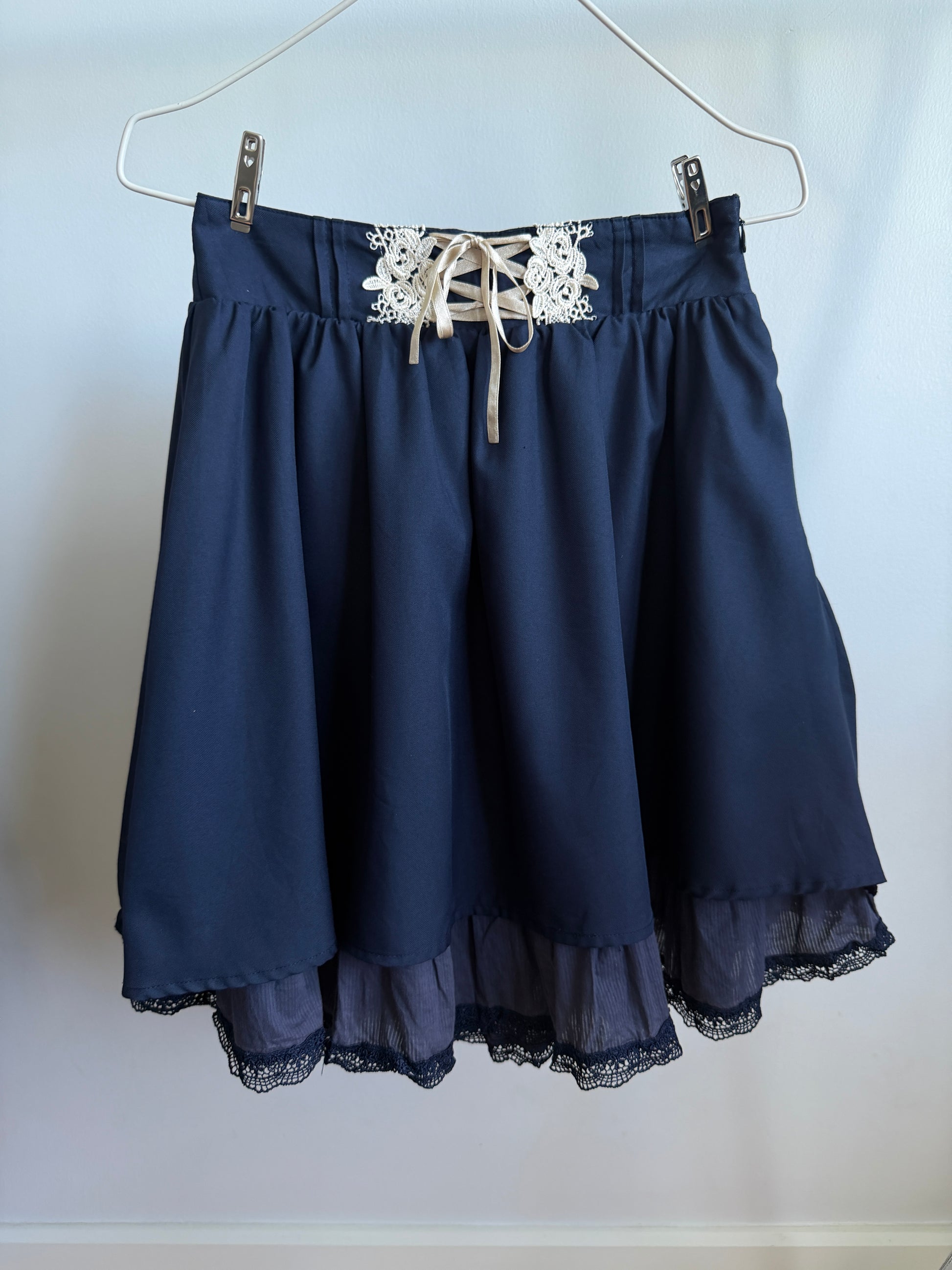 A navy blue skirt with a lace-up waistband and white floral lace detail, hanging from a metal hanger against a plain light background.