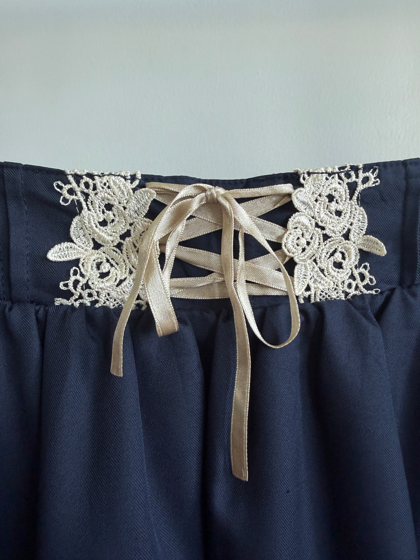 Close-up of a navy blue skirt with white floral lace applique and beige ribbon laces at the waistband.