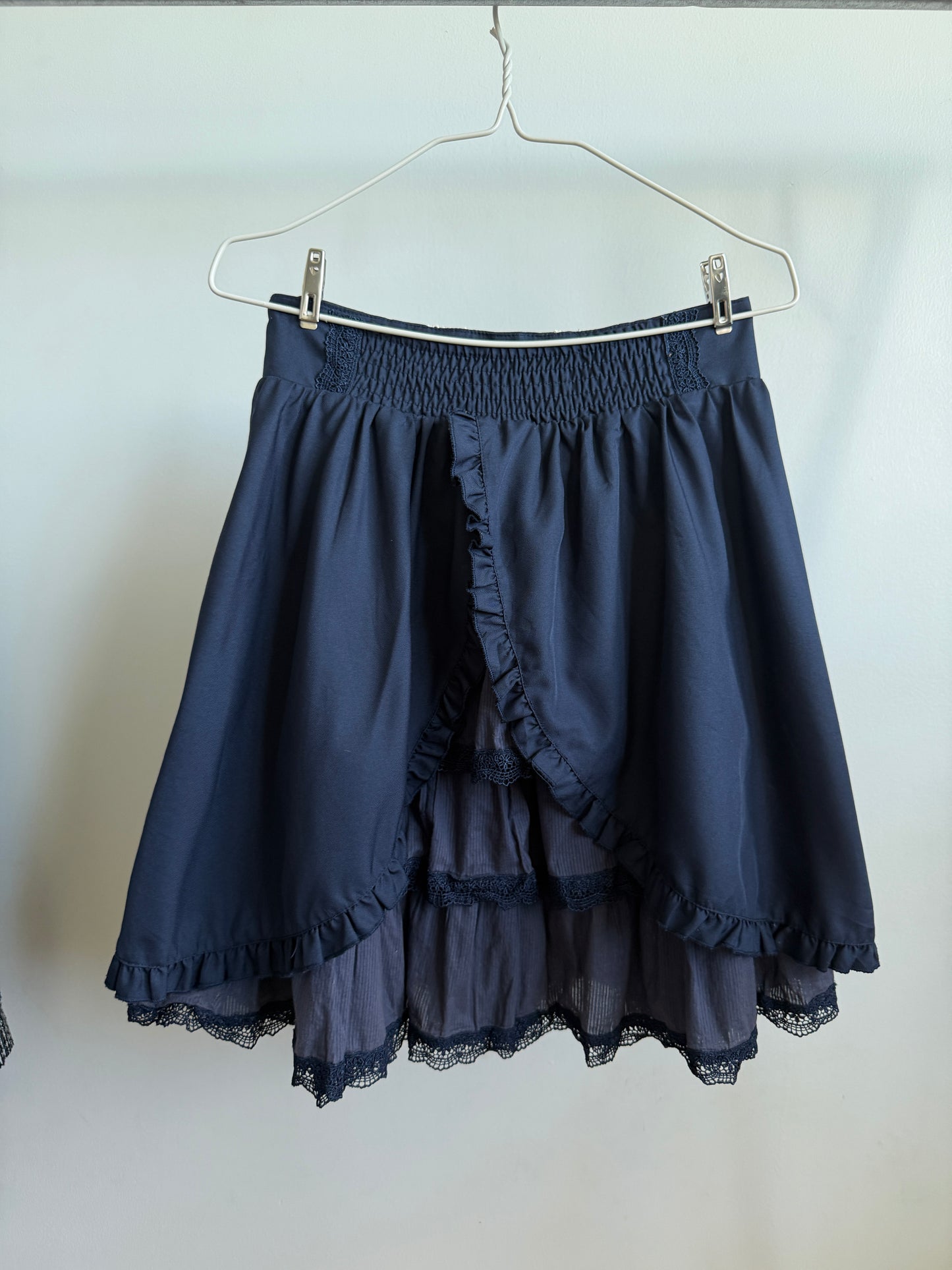 A dark navy blue high-low skirt with a smocked elastic waistband, ruffled front overlay, and layered sheer tulle skirt with lace trim, hanging on a metal hanger against a plain light background.