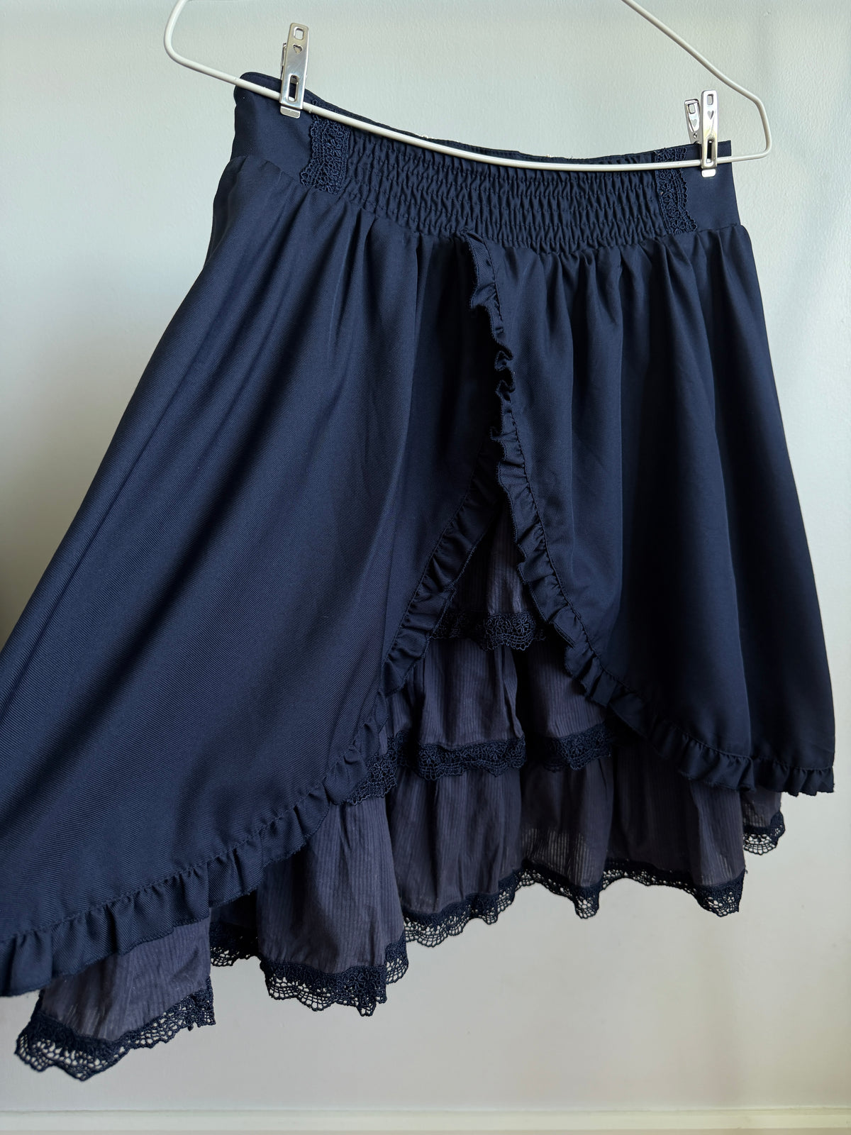 A dark navy blue high-low skirt with a smocked waistband, layered ruffle details, and lace trim, hanging on a metal hanger against a plain light gray wall.