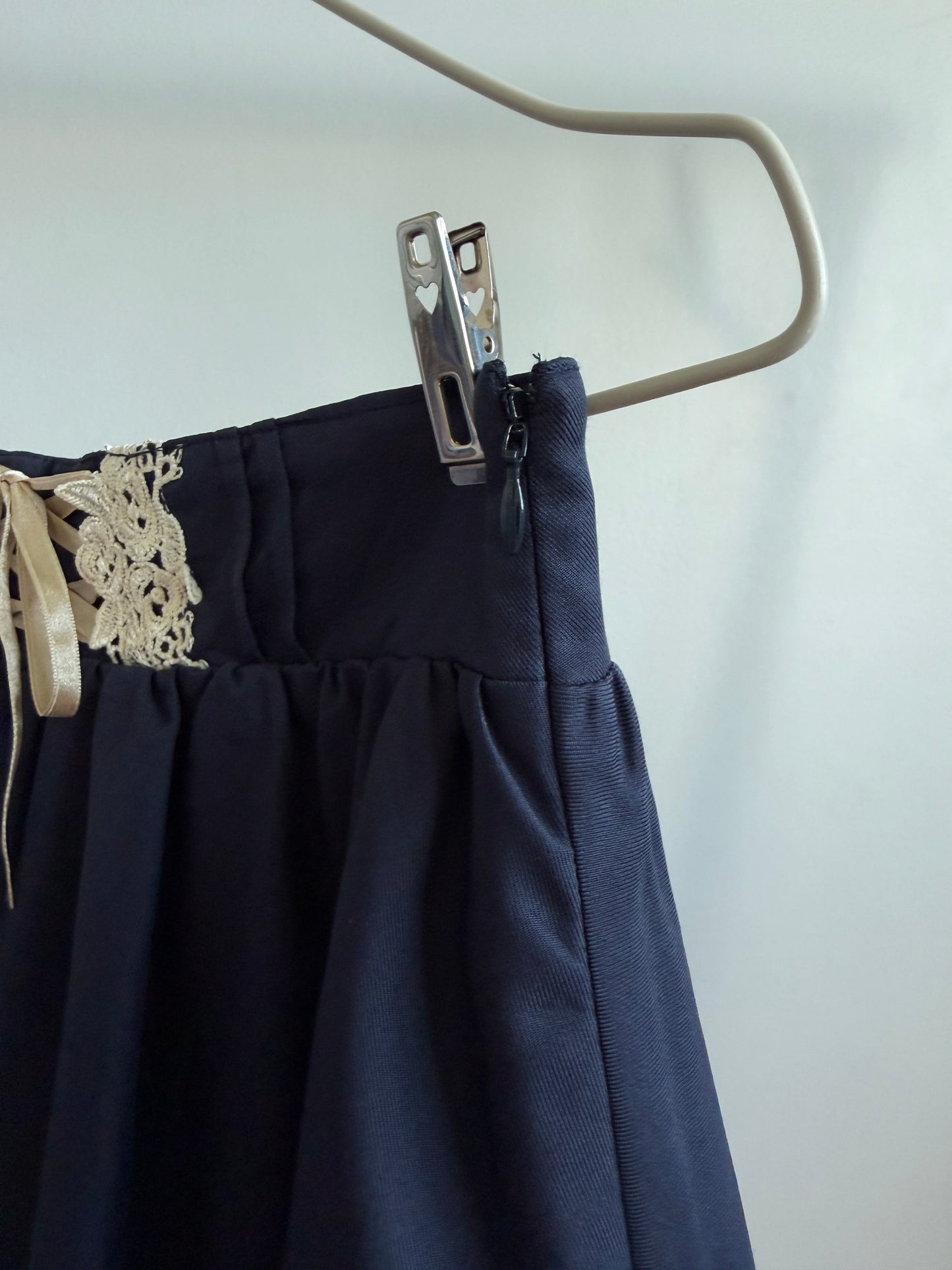 A close-up of a dark navy pleated skirt hanging from a beige hanger with a metal clothespin. The skirt features a gathered waistband, a decorative beige lace applique on the left side, and a matching beige ribbon tie.