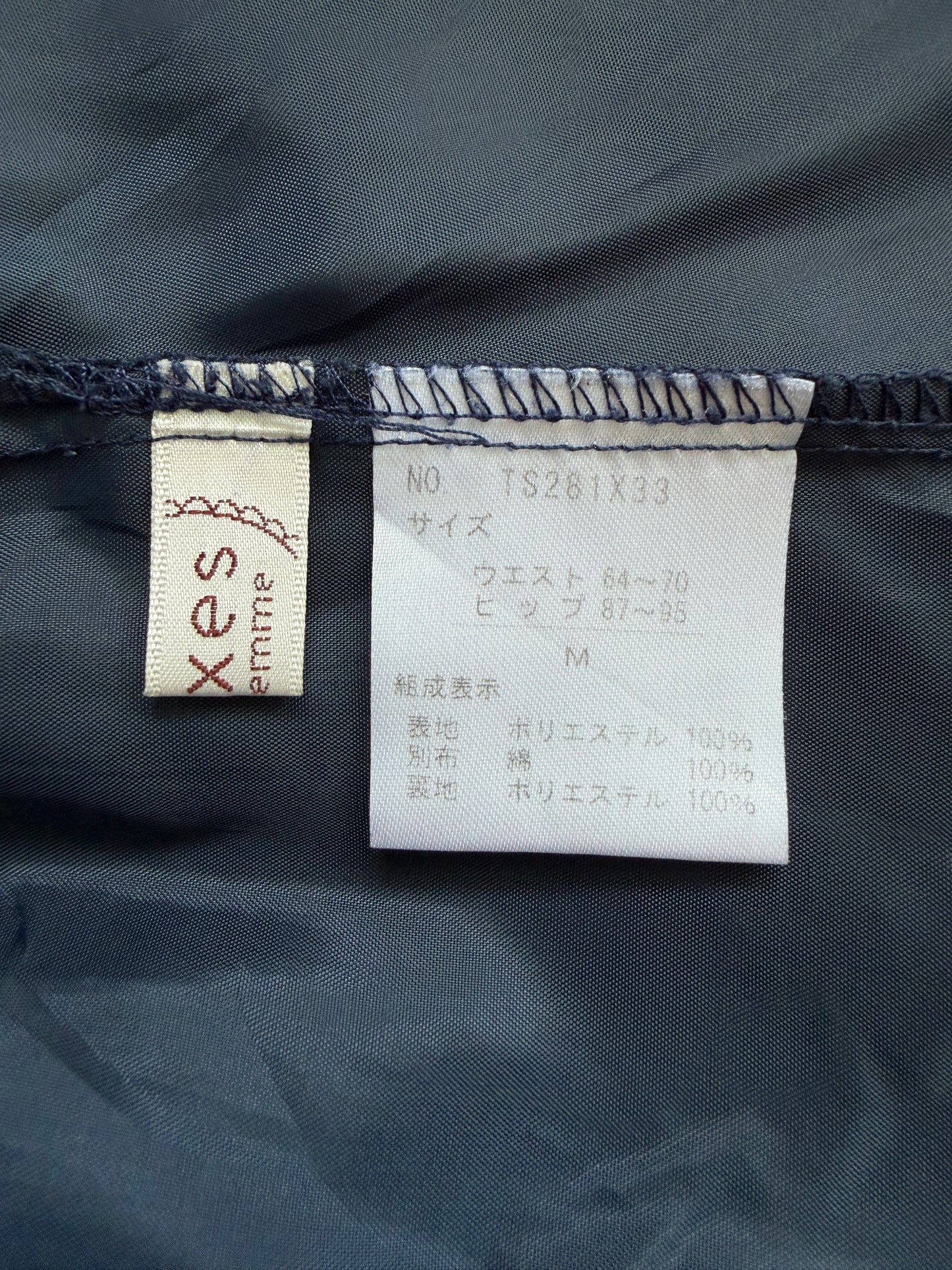 Close-up of a garment's interior labels showing the brand 'xes entrie' on a beige tag and size information on a white tag, set against dark navy blue fabric.