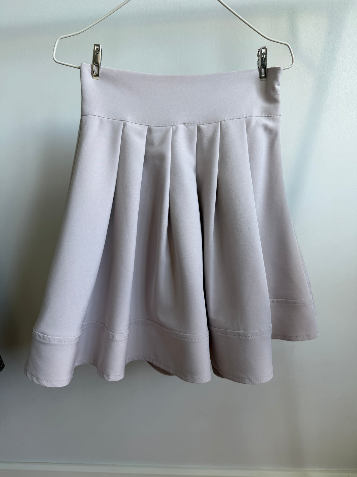 A light gray pleated skirt hanging from a metal hanger against a plain light gray wall.