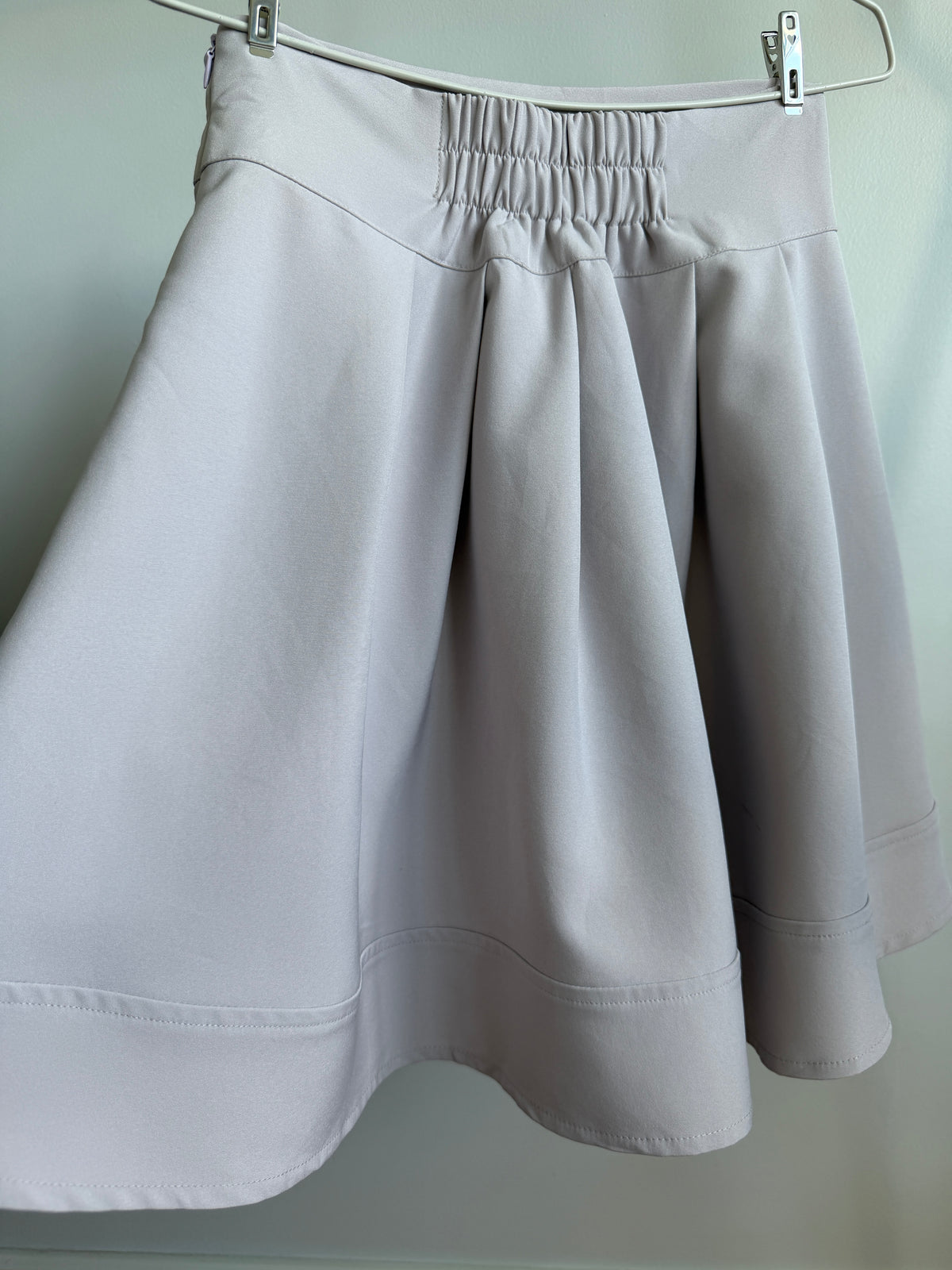 A light gray pleated A-line skirt with an elastic waistband, hanging from a metal hanger against a plain light gray wall.