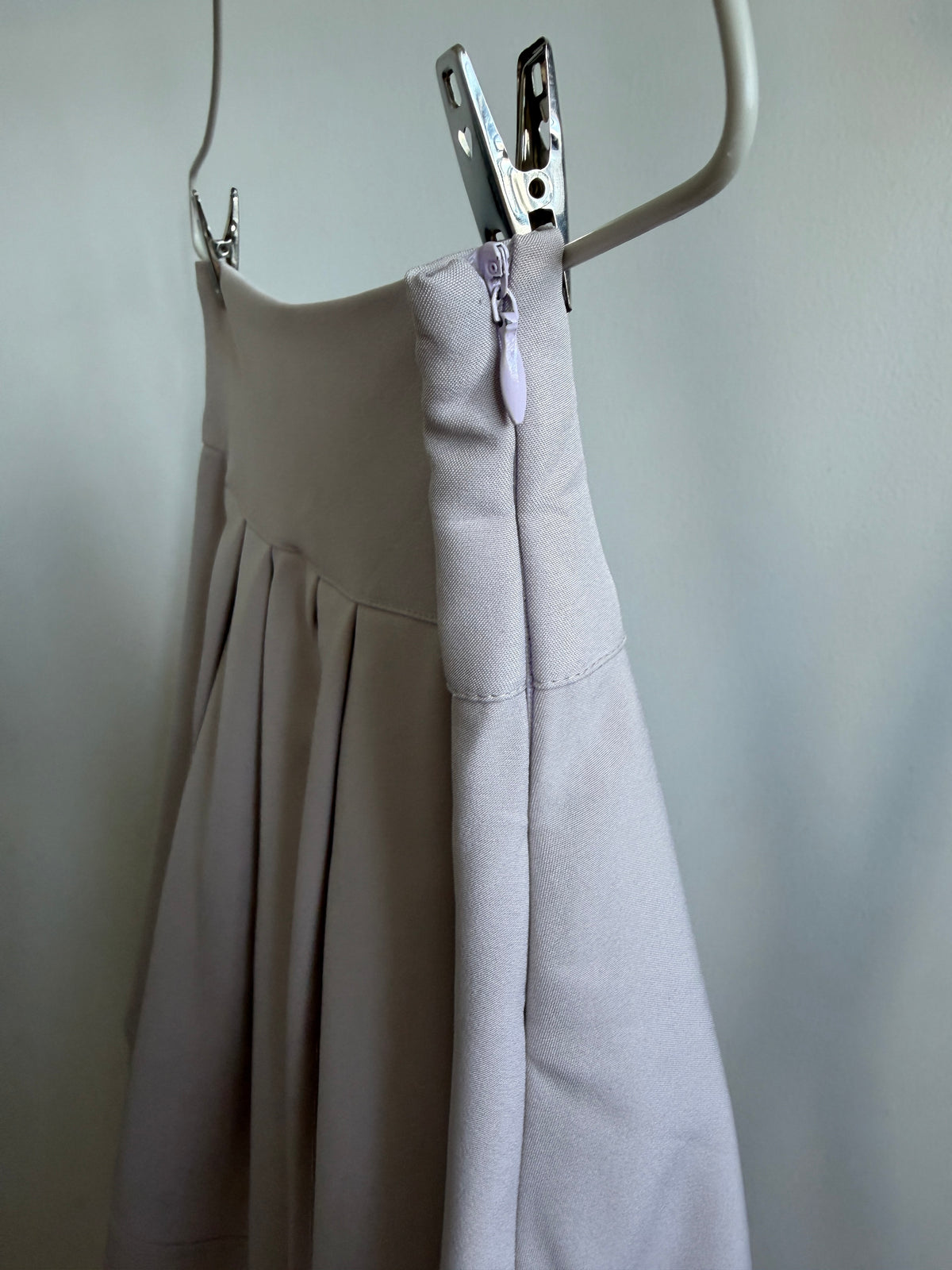 A close-up of a light beige pleated skirt or dress hanging from a white hanger, with a metal zipper and silver zipper pull clipped at the waist. The fabric is shown against a plain light gray wall.