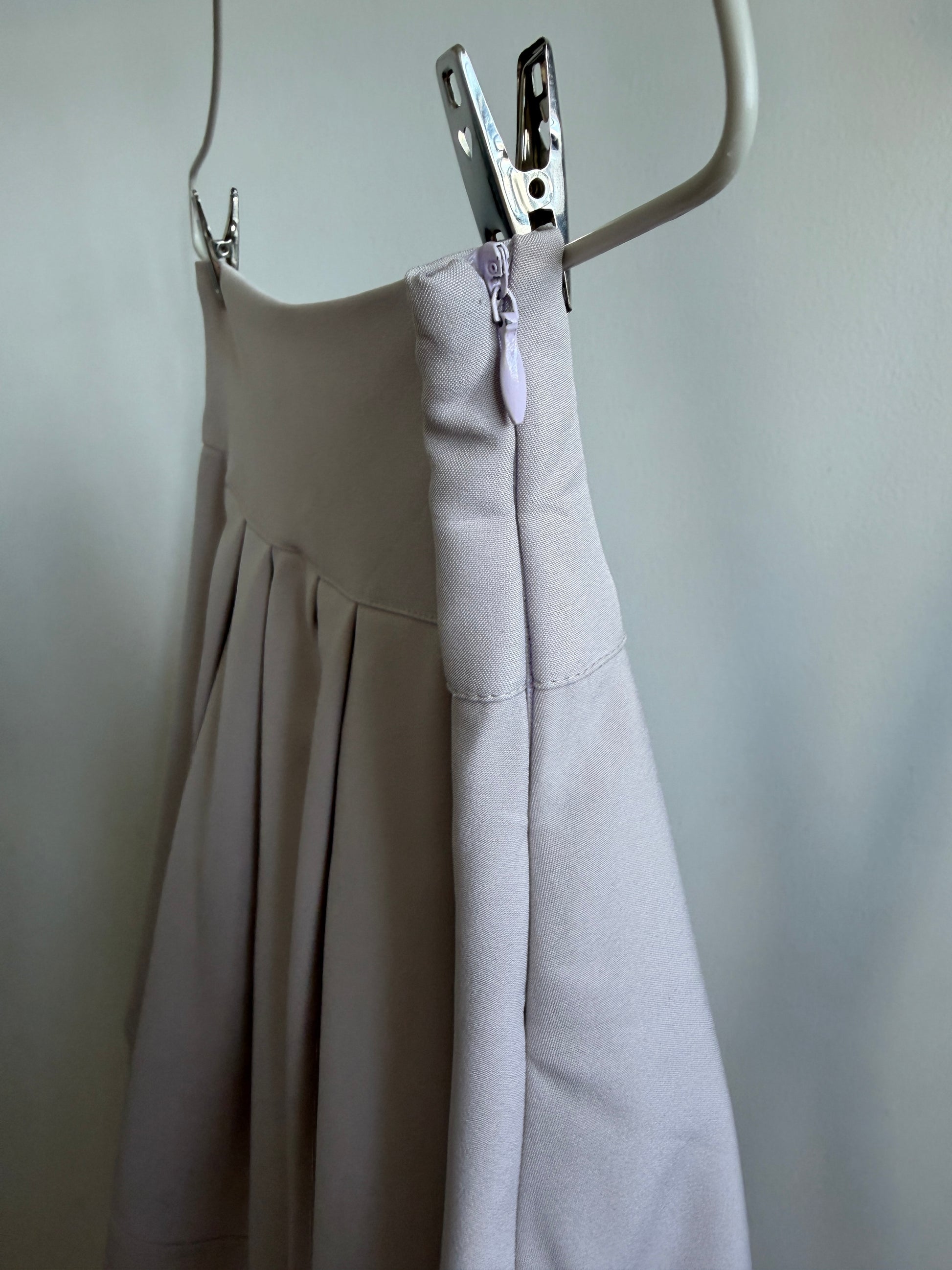A close-up of a light beige pleated skirt or dress hanging from a white hanger, with a metal zipper and silver zipper pull clipped at the waist. The fabric is shown against a plain light gray wall.