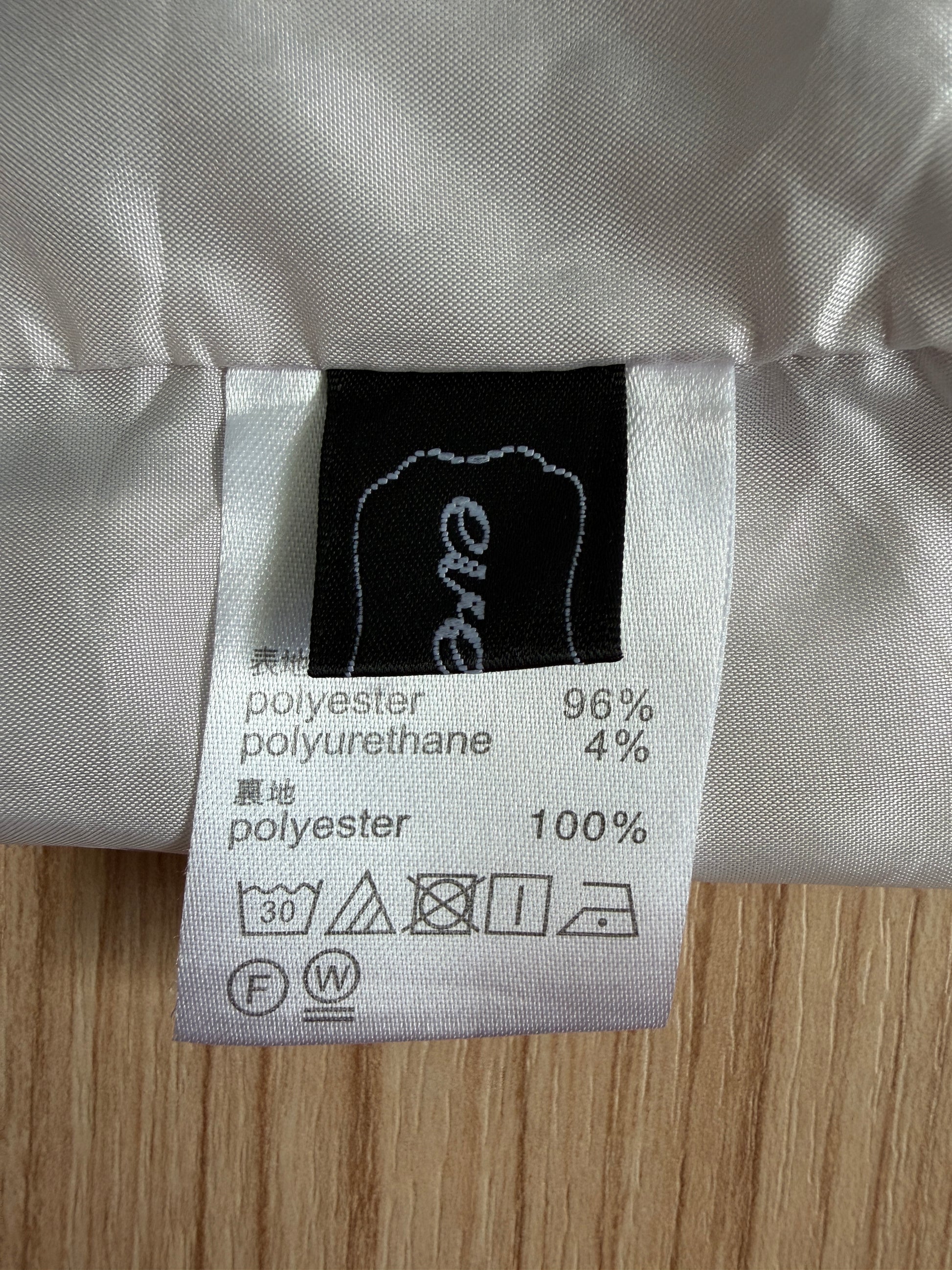 Close-up of a garment's interior care label showing the brand 'eve' on a black tag, with polyester and polyurethane content details and care symbols, laid flat on a light wood surface.