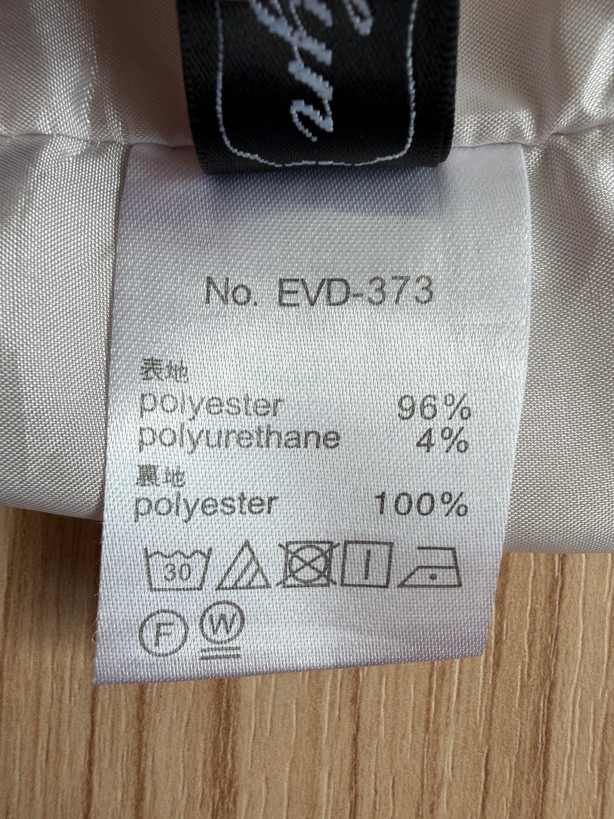Close-up of a garment's interior care label showing composition and care instructions, with a black neck label partially visible above it on a light fabric surface.