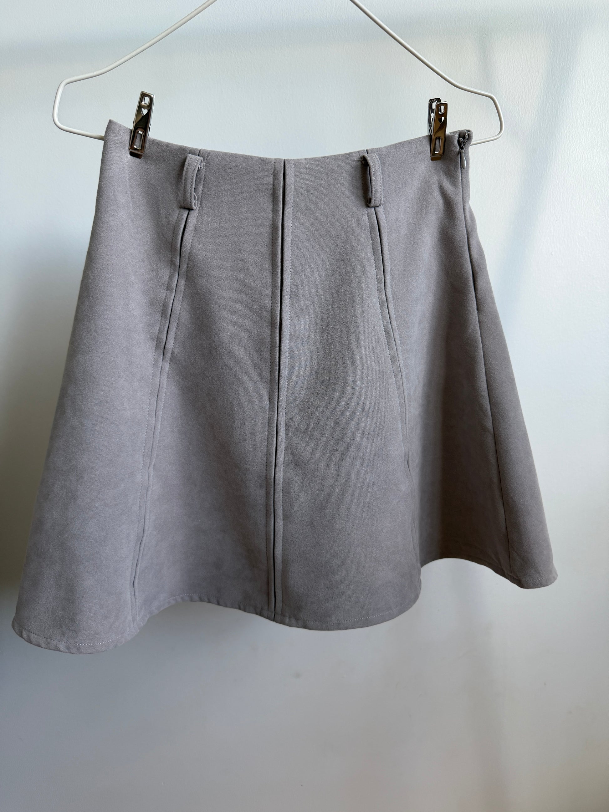 A light gray A-line skirt made of textured faux suede, hanging from a metal hanger with two clips against a plain white wall.