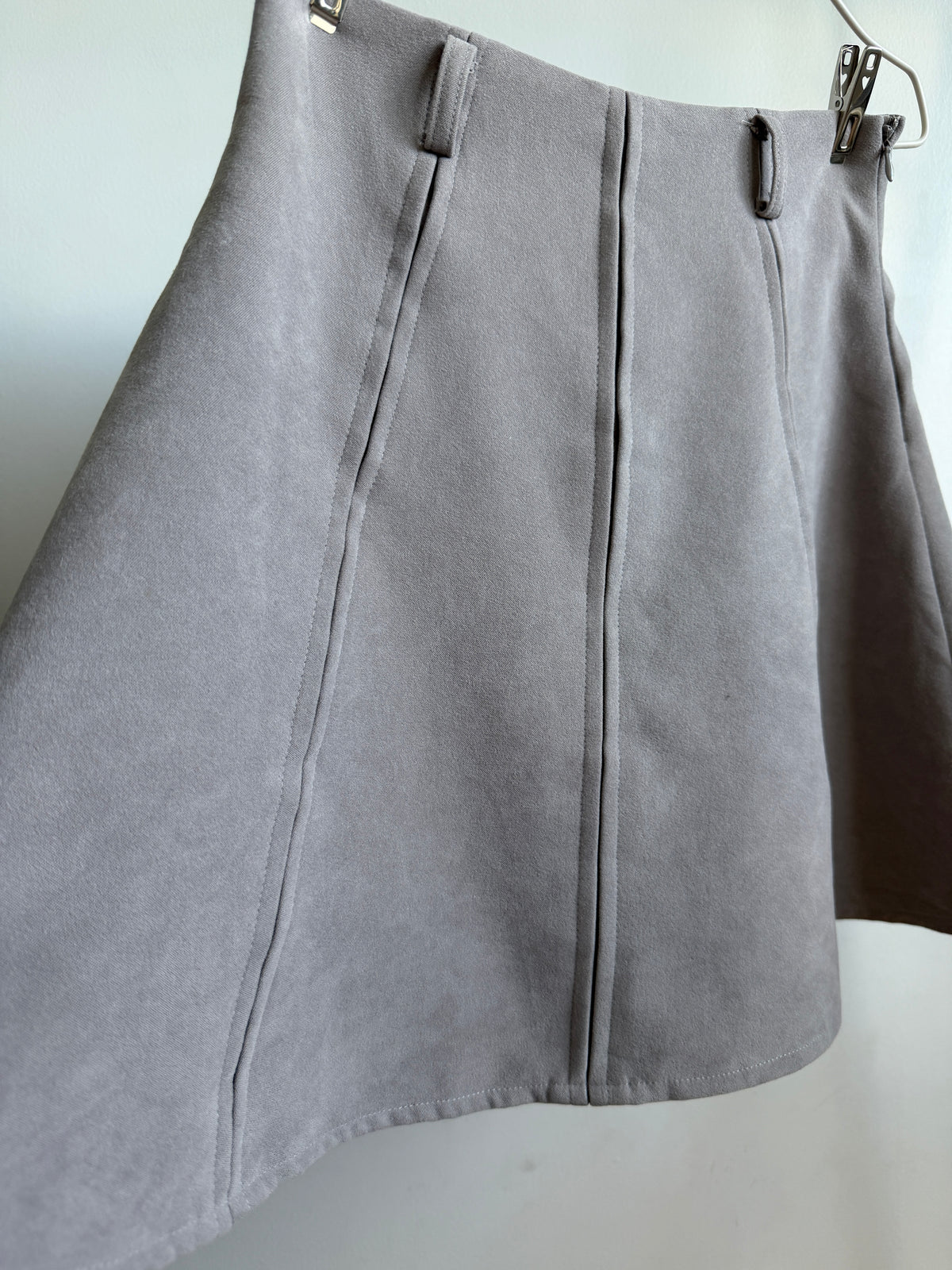 A light gray suede skirt with vertical seam detailing, hanging from a metal hanger against a plain light background.