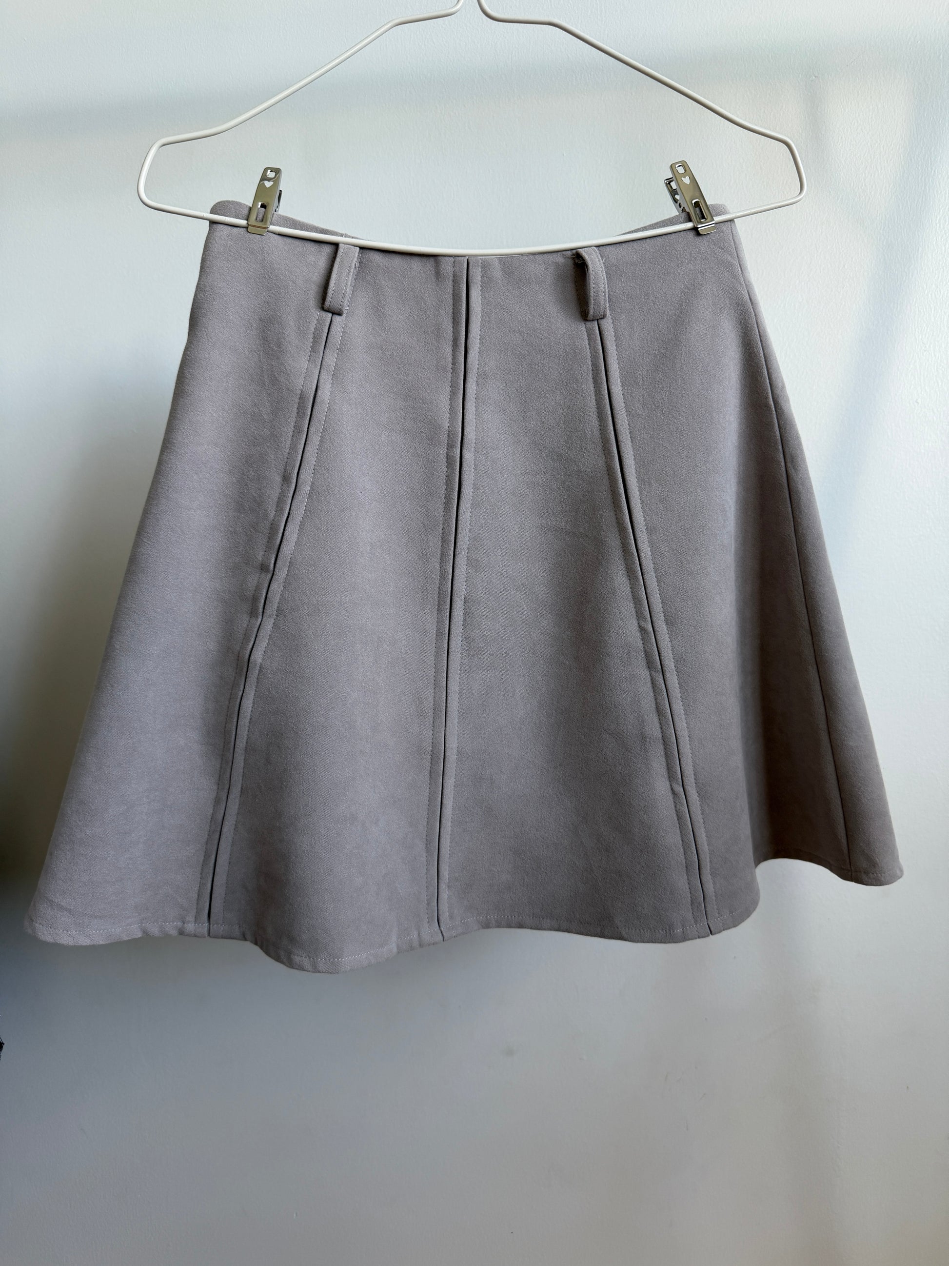 A light gray pleated A-line skirt hanging from a white wire hanger against a plain light gray wall.