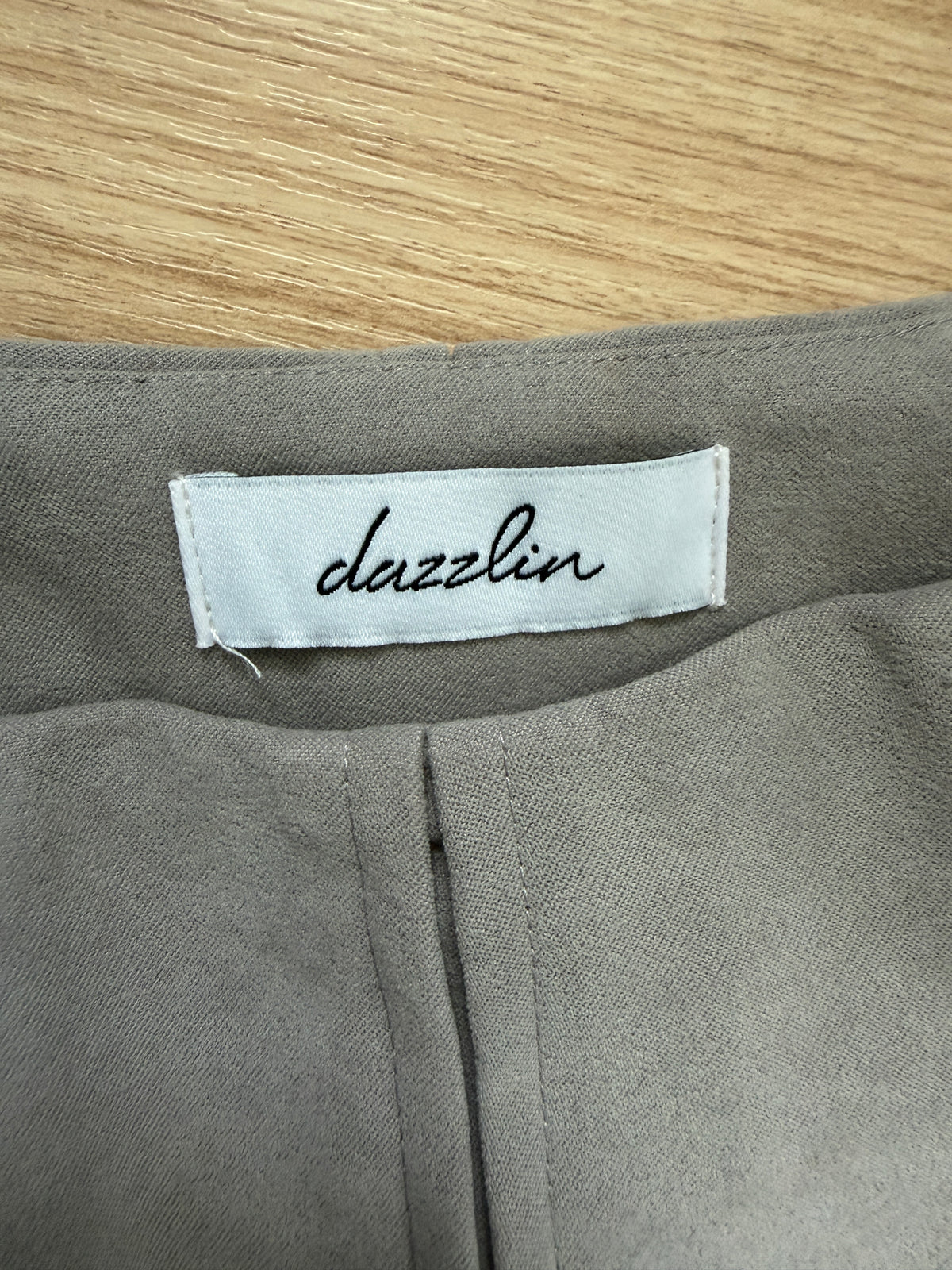 Close-up of a gray fabric garment showing a white sewn-in label with the brand name 'dazzlin' in black script, placed on a light wood surface.