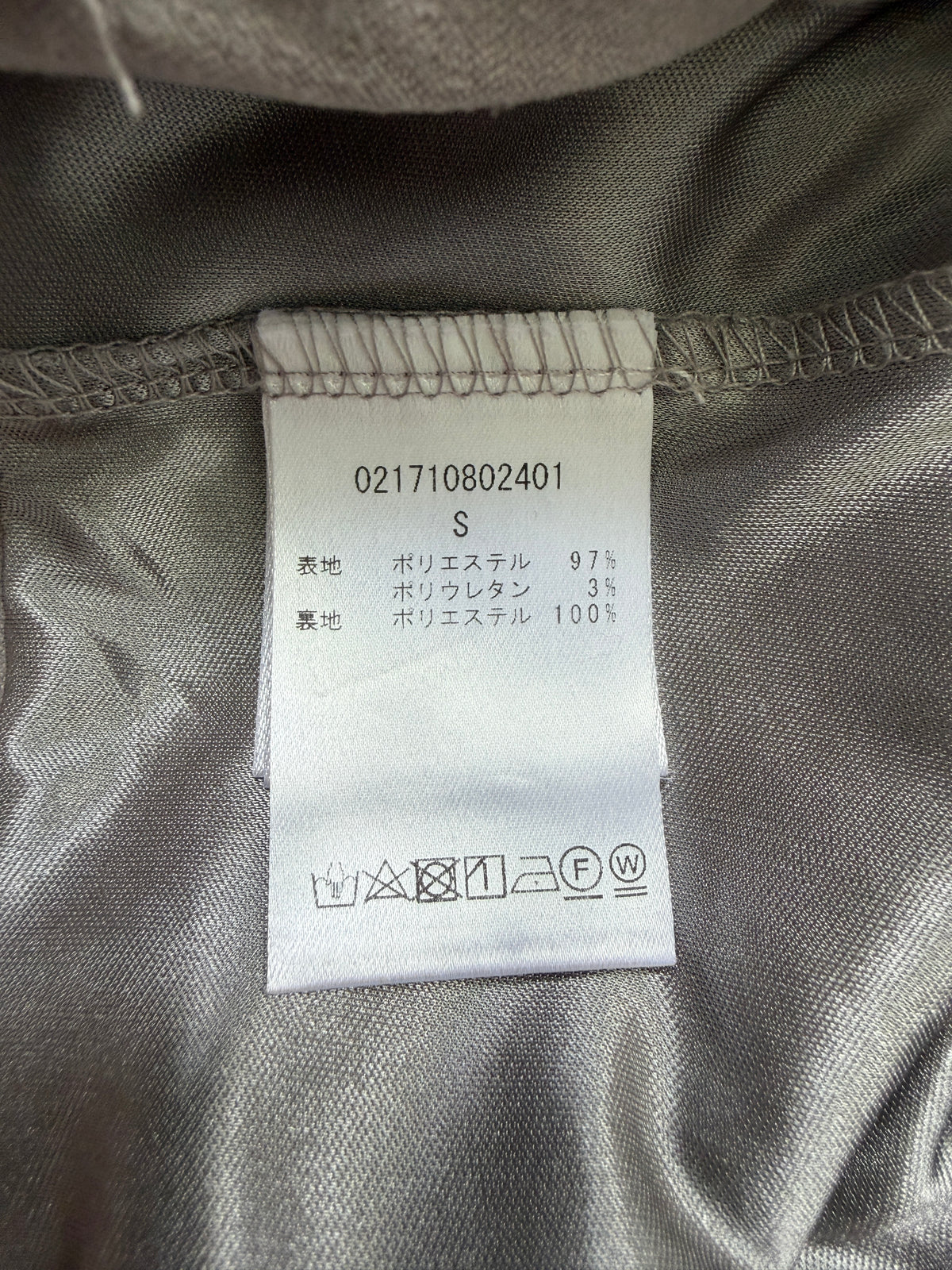 Close-up of a garment's interior neck label showing size S and fabric composition in Japanese, set against a gray satin-like lining.