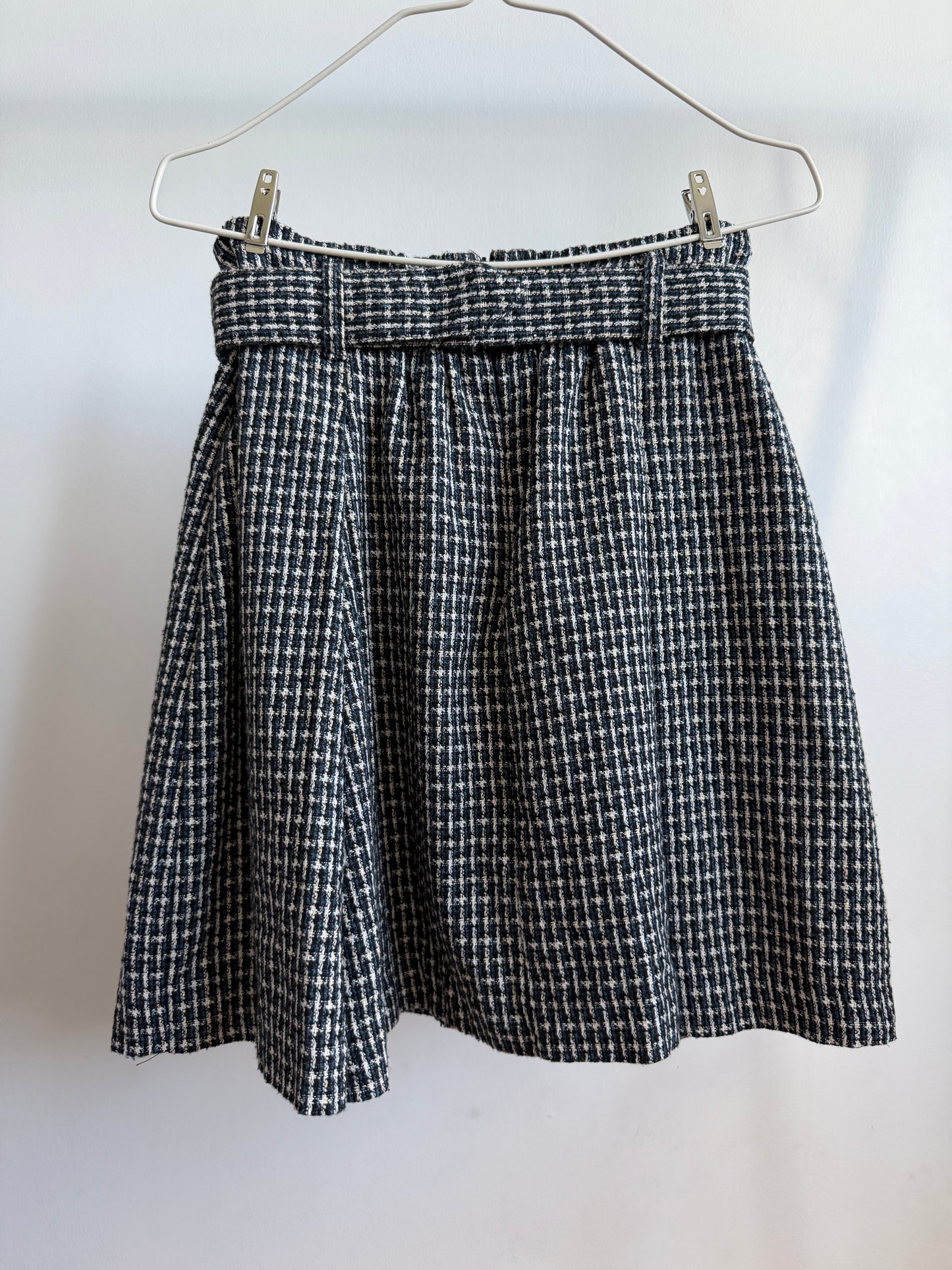 A black and white houndstooth patterned skirt hanging from a metal hanger against a plain white wall.