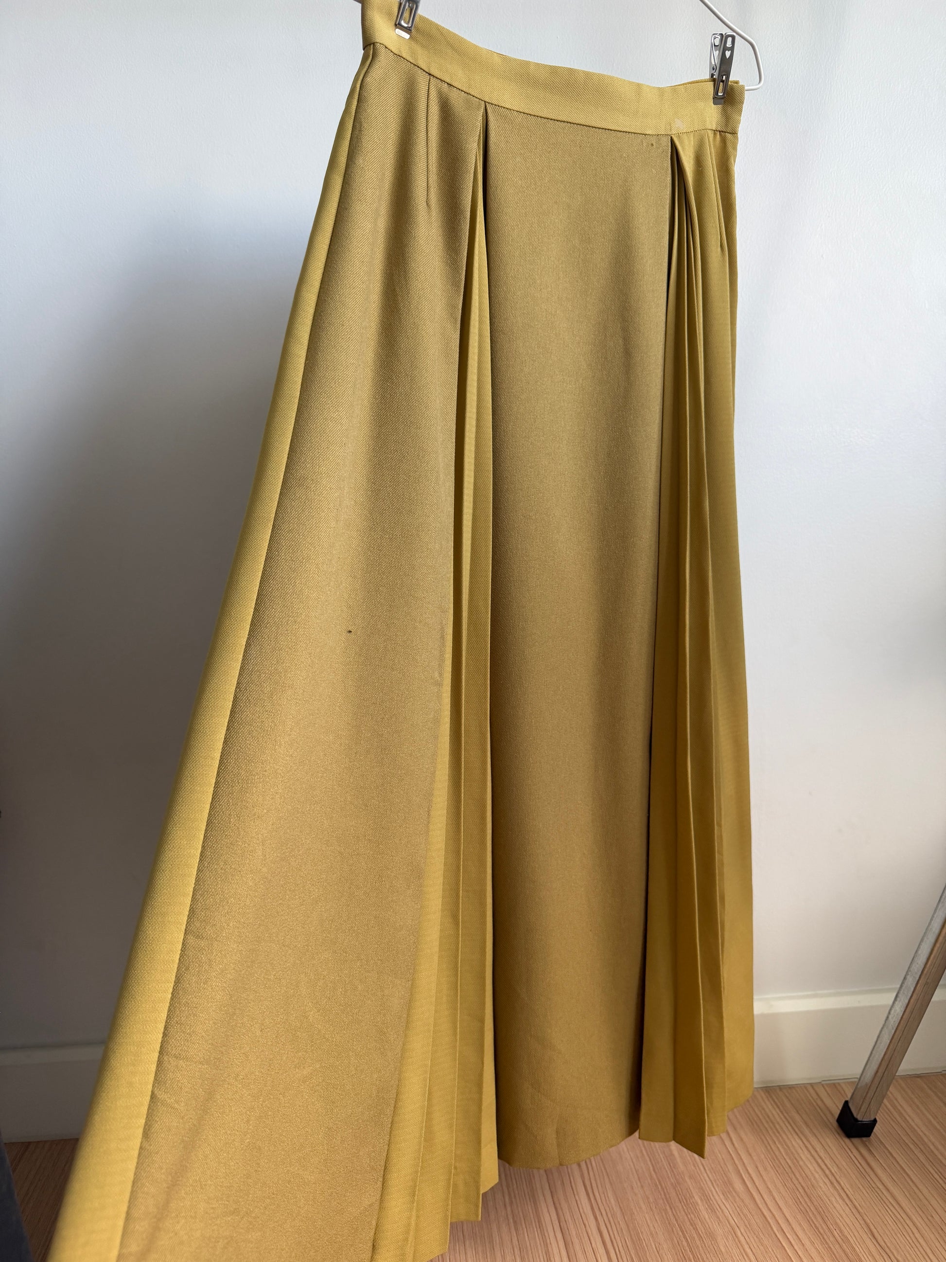 A pleated midi skirt in a mustard yellow color, hanging from a metal hanger against a plain white wall.