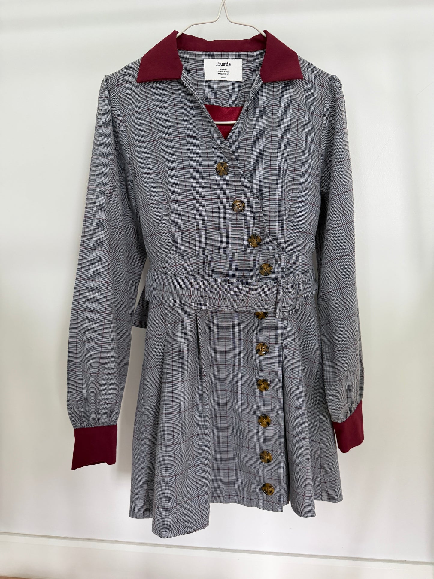 A gray plaid belted coat dress with a double-breasted wrap front, brown buttons, and contrasting burgundy collar, cuffs, and lining, hanging on a white hanger against a white wall.