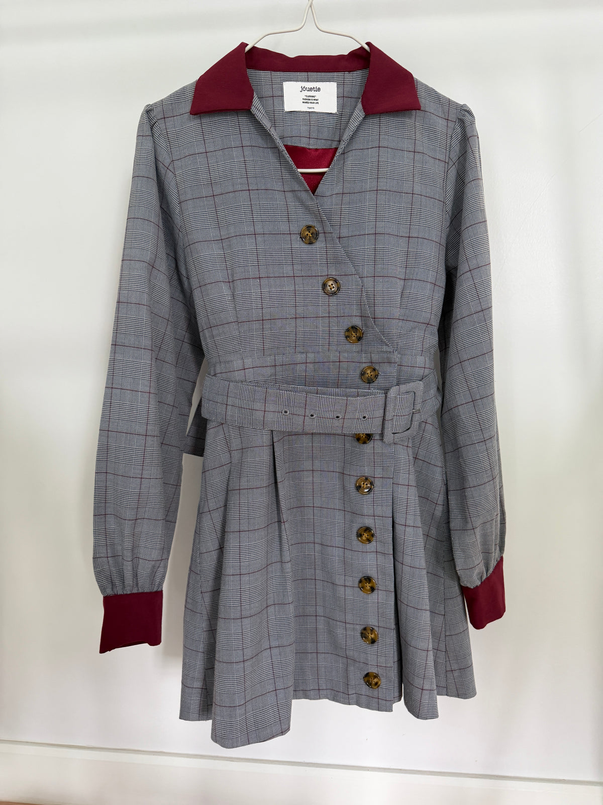A gray plaid belted coat dress with a double-breasted wrap front, brown buttons, and contrasting burgundy collar, cuffs, and lining, hanging on a white hanger against a white wall.