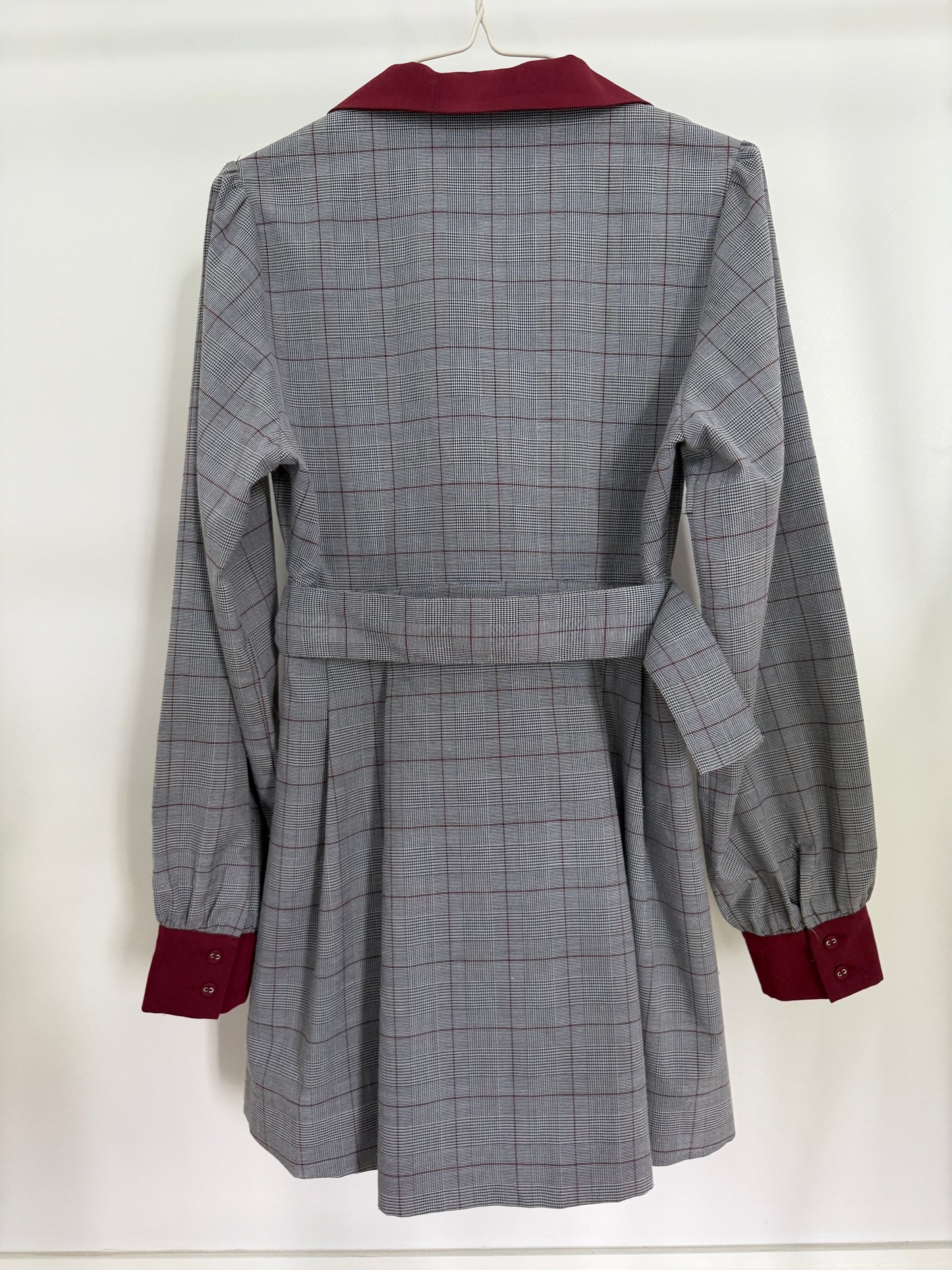 A gray and white plaid belted coat with contrasting dark red collar, cuffs, and button details, hanging on a white hanger against a white background.